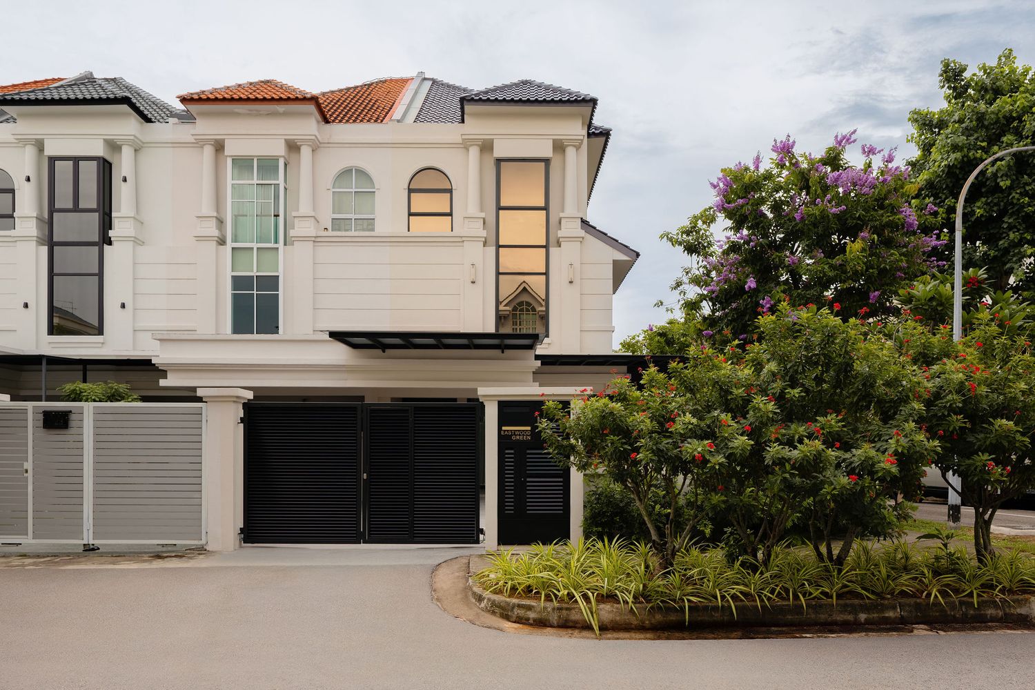 White two-story colonial house with black garage doors, lush green trees, and a curved driveway in a suburban neighborhood.