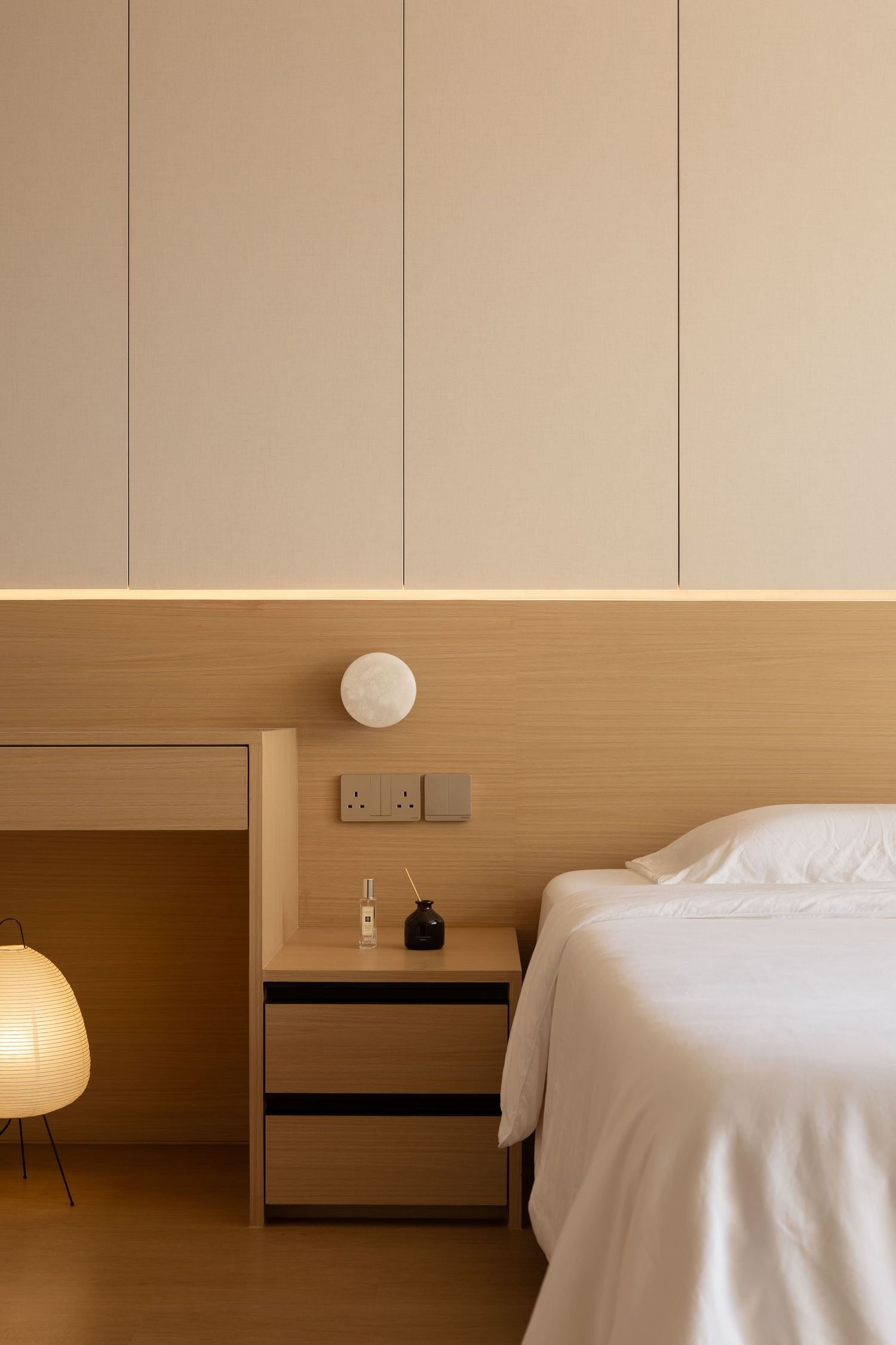 A minimalist hotel room interior with light wood paneling and white bedding in warm beige tones.
