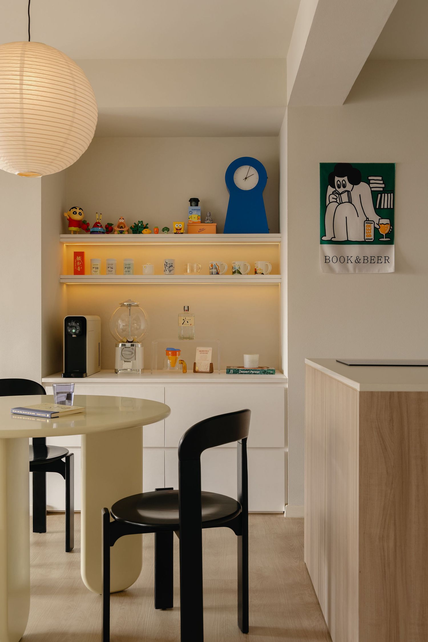 Modern kitchen nook with built-in shelves, blue clock, colorful decor, black chairs, round table, and a Book & Beer poster on wall.