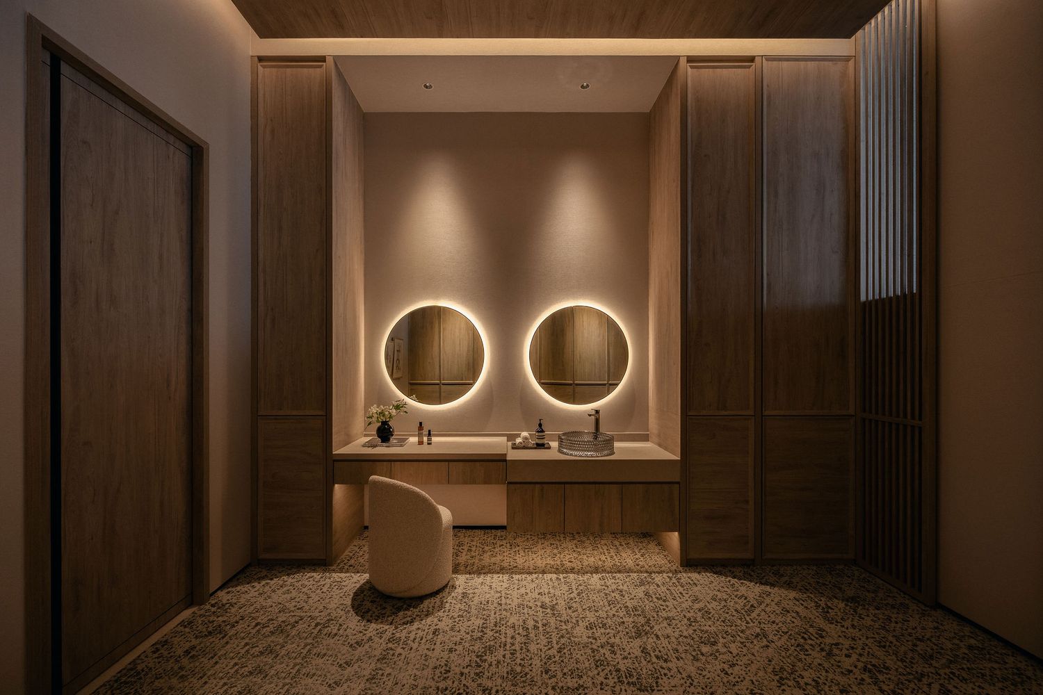Elegant bathroom vanity with dual round backlit mirrors, warm wood paneling, and a modern white chair on textured flooring.