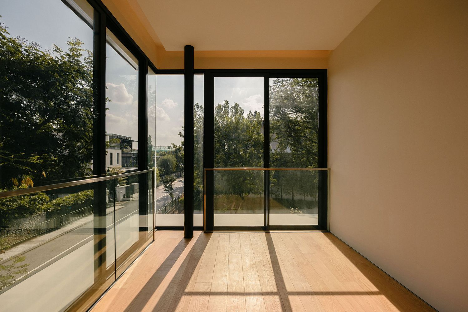 Sunlit modern balcony room with floor-to-ceiling glass walls, wooden flooring, and a glass railing overlooking green trees.