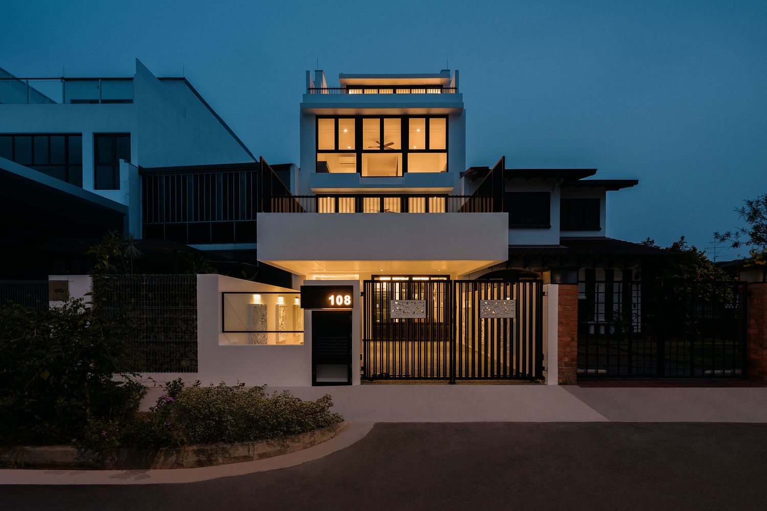 Modern multi-storey house with illuminated windows, rooftop terrace and gated entrance, photographed at dusk with a deep blue sky.