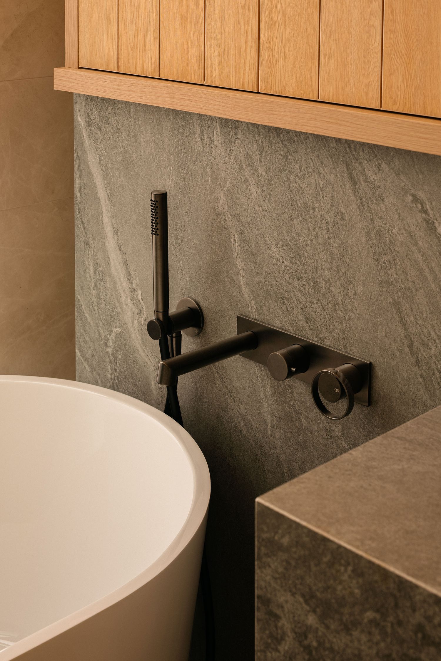 Matte black wall-mounted bathtub faucet with handheld shower on a gray slate wall beside a white freestanding tub and oak cabinetry.