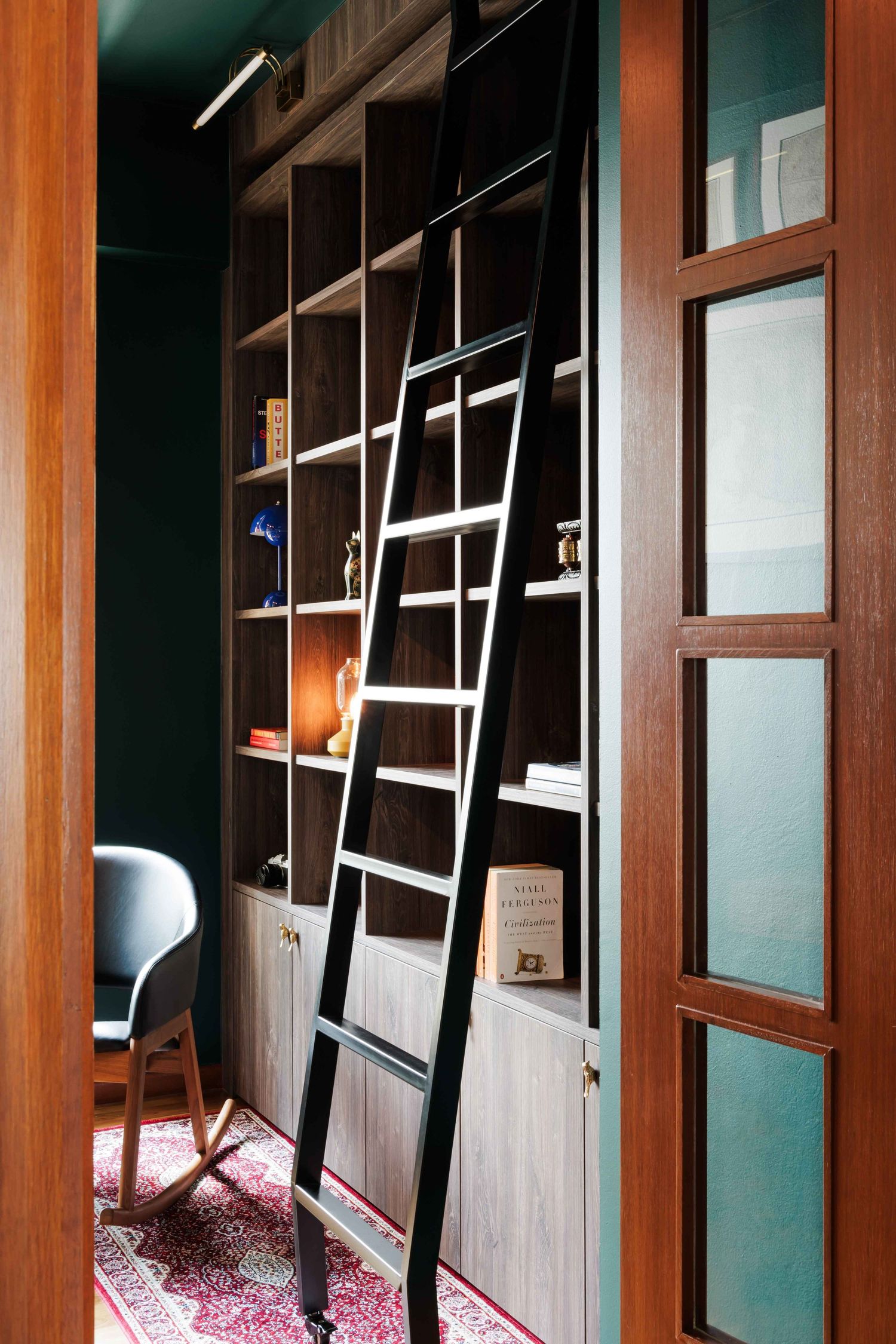 Modern home library with tall wooden shelving unit featuring a sleek black ladder and oriental rug on the floor.