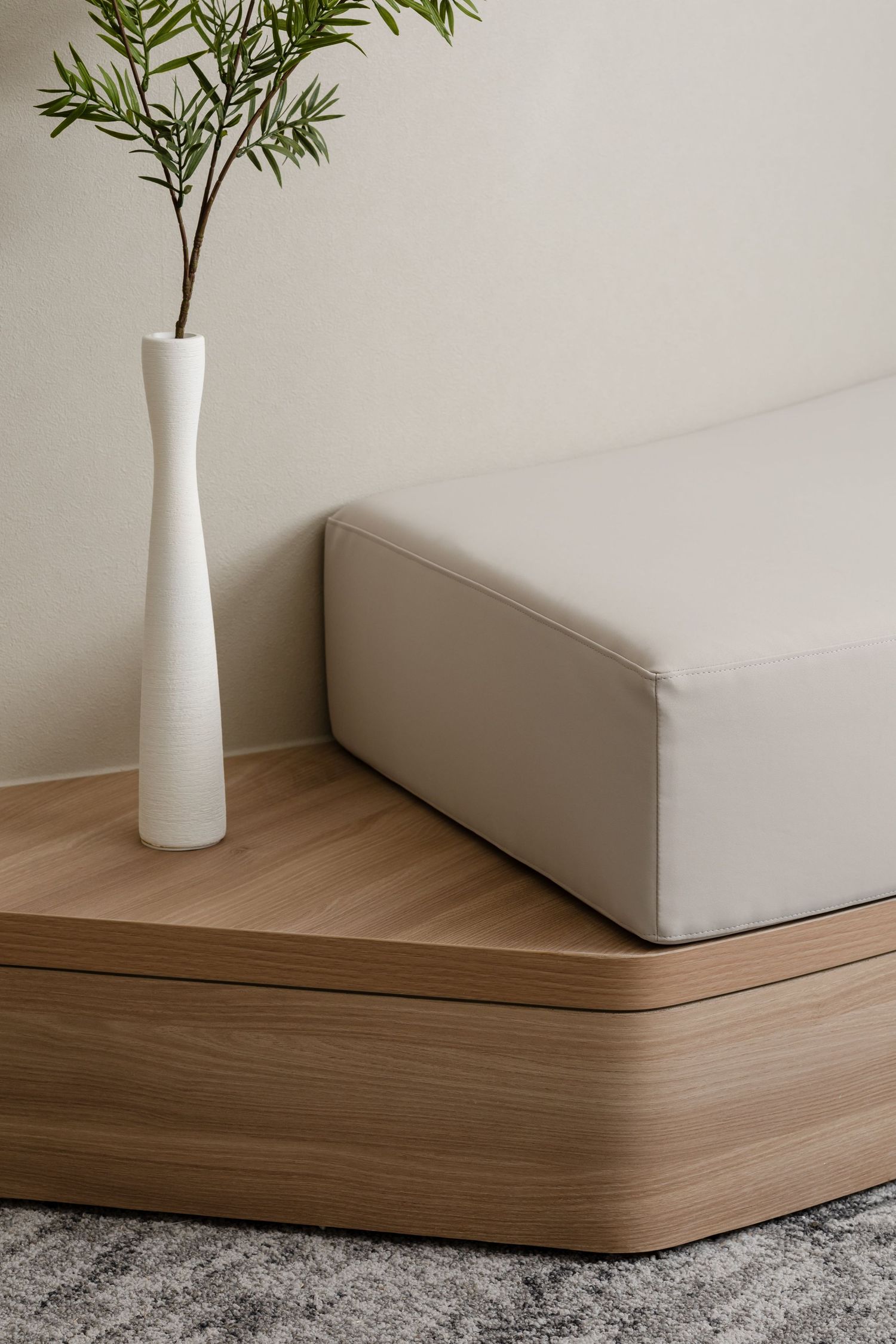Modern minimalist interior with tall white vase holding green plant stems on wooden platform with beige cushioned seating.