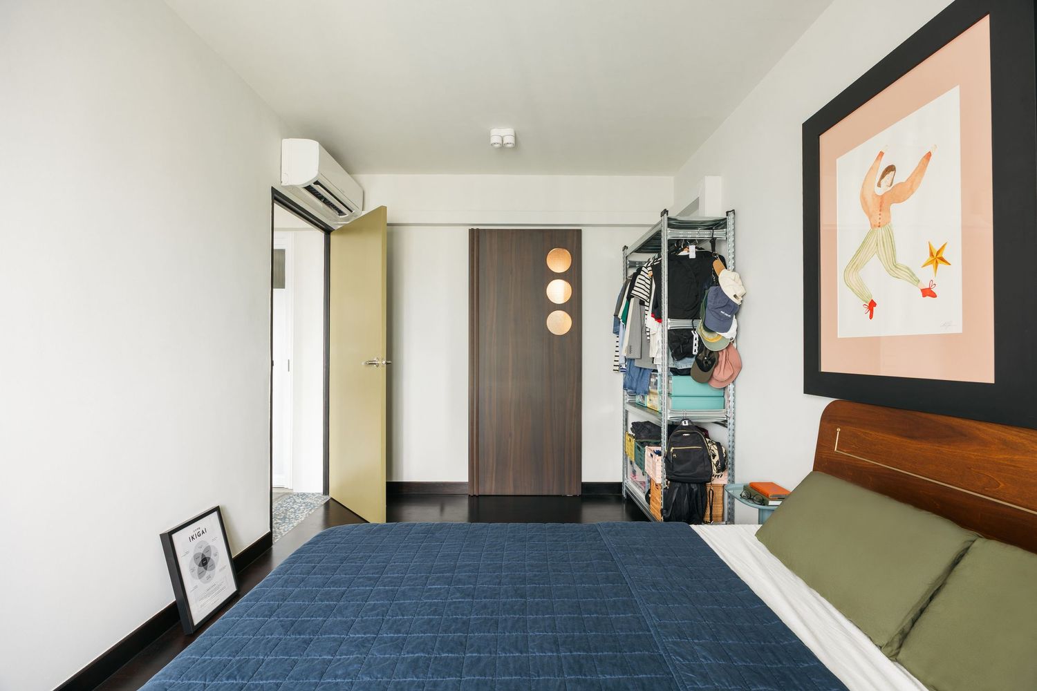 Modern bedroom with blue bedspread, wooden wardrobe, open shelving unit, wall art, and split air conditioning unit.