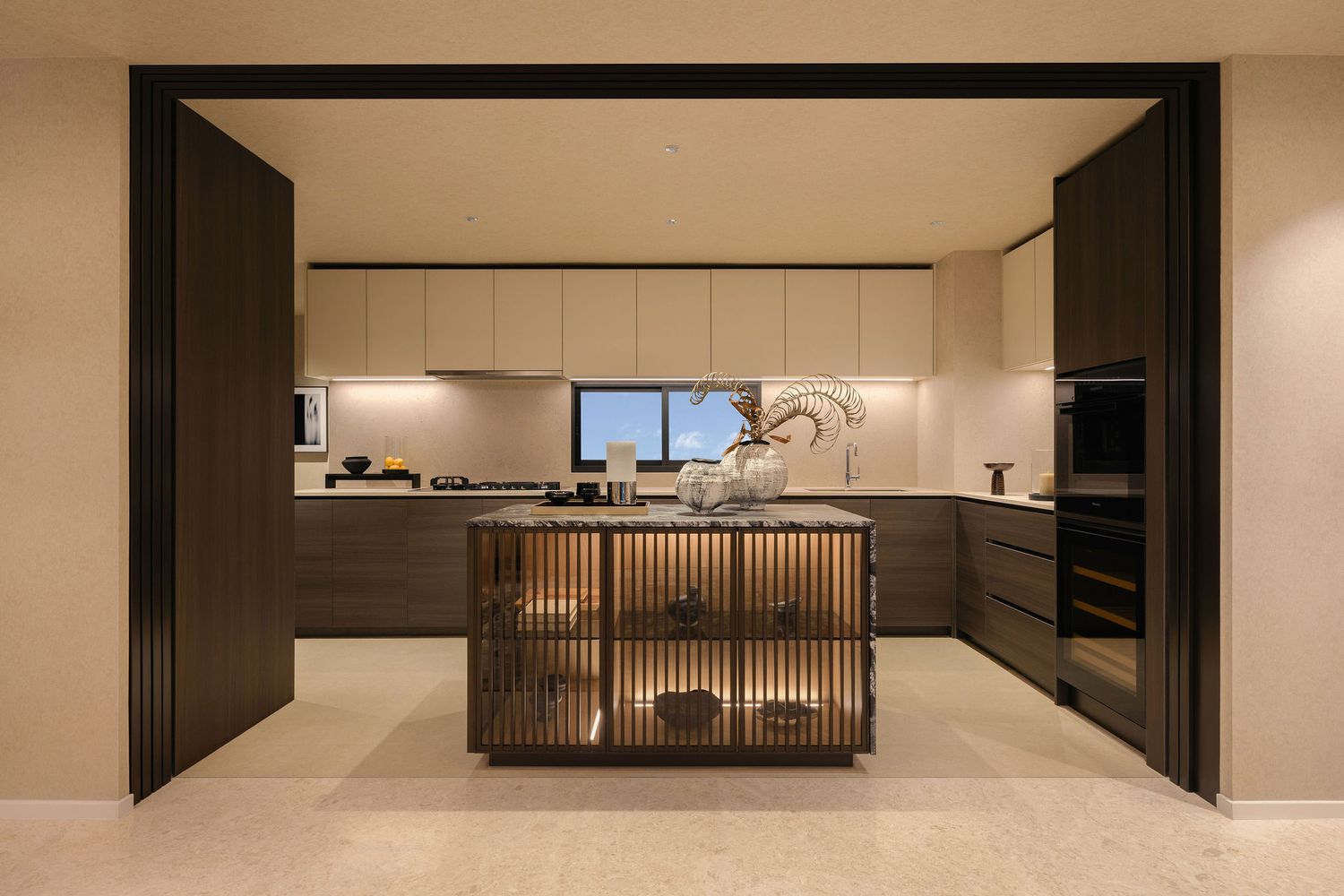 Modern luxury kitchen with a central island with built-in shelving, dark wood cabinetry, and built-in ovens viewed through a sliding door.
