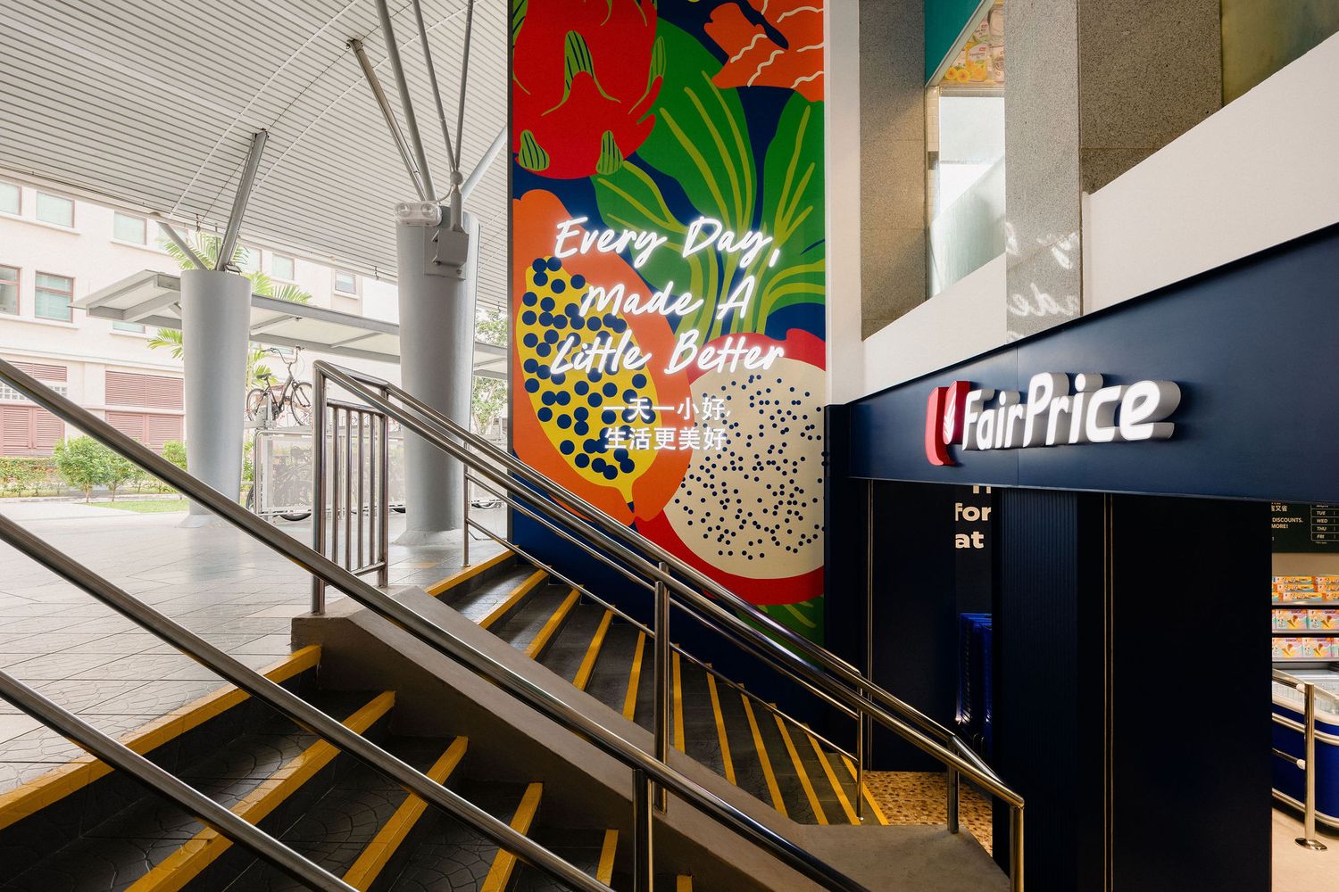 Colorful tropical fruit mural reading Every Day Made a Little Better beside a staircase leading to a FairPrice supermarket entrance.
