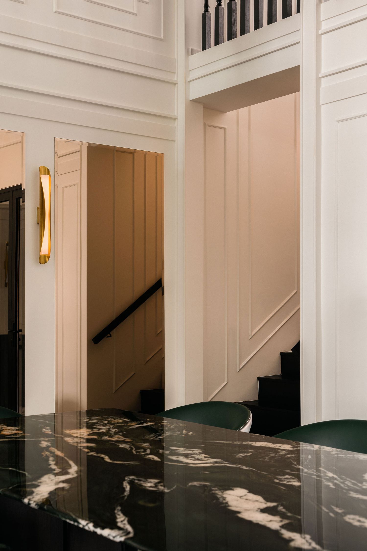Elegant foyer with white paneled walls, a brass wall sconce, black marble table with white veining, green chairs, and a staircase beyond.