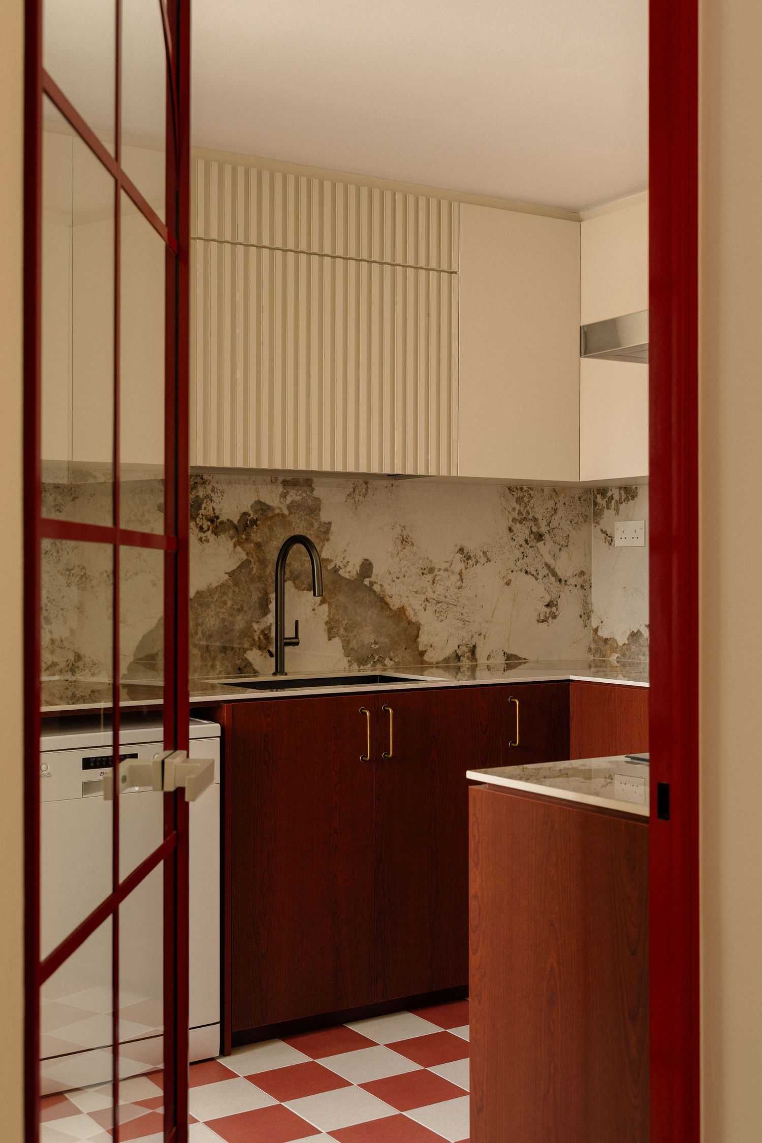 Modern kitchen with red checkered tile floor, dark red wood cabinets, black faucet, fluted upper cabinets and a marble backsplash.