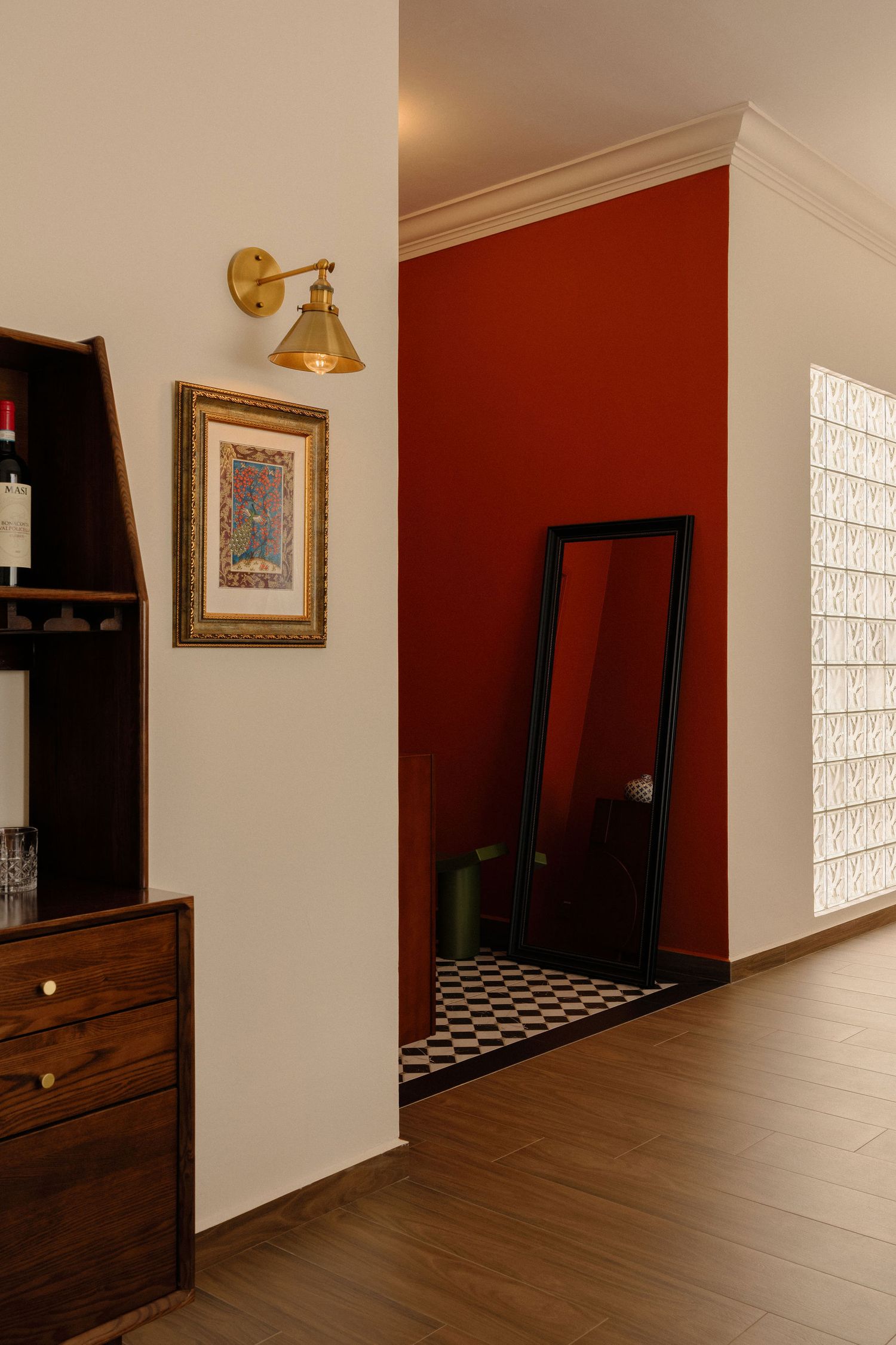 Warm hallway with a deep red accent wall, brass wall sconce, framed art, wooden bar cabinet, and a floor mirror over checkered tile.