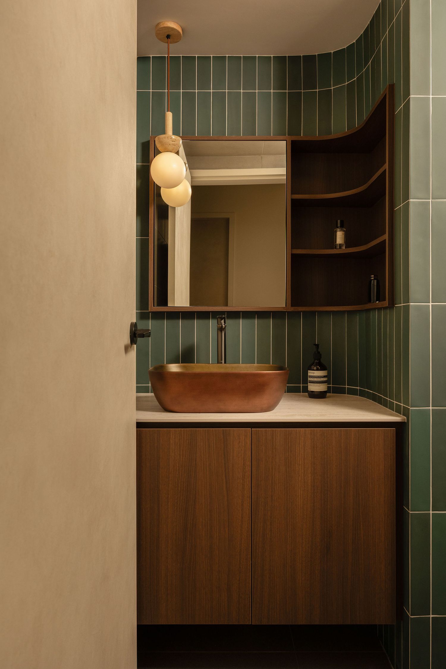 Modern powder room with green vertical tiles, a copper vessel sink, walnut vanity, built-in corner shelves, and a globe pendant light.