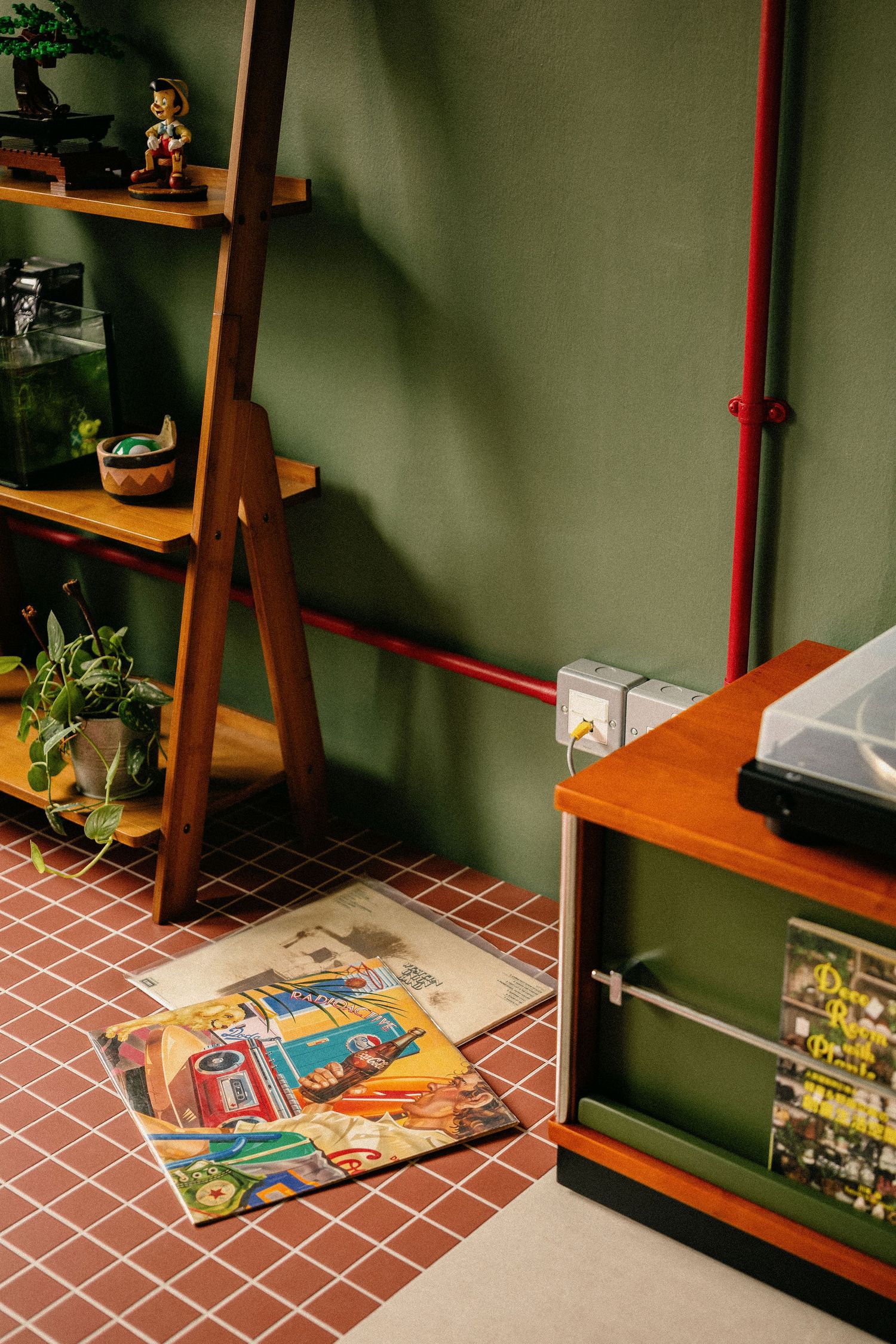 Cozy green-walled room with a wooden ladder shelf, potted plants, and vinyl records on a red tile floor beside a turntable stand.