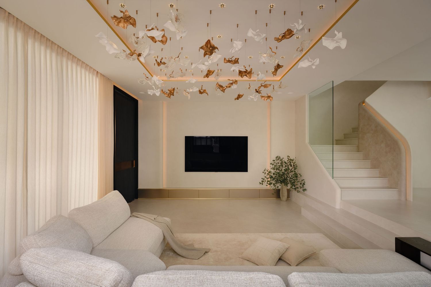 Modern minimalist living room with white sofa, gold floral ceiling installation, wall-mounted TV, and elegant staircase.
