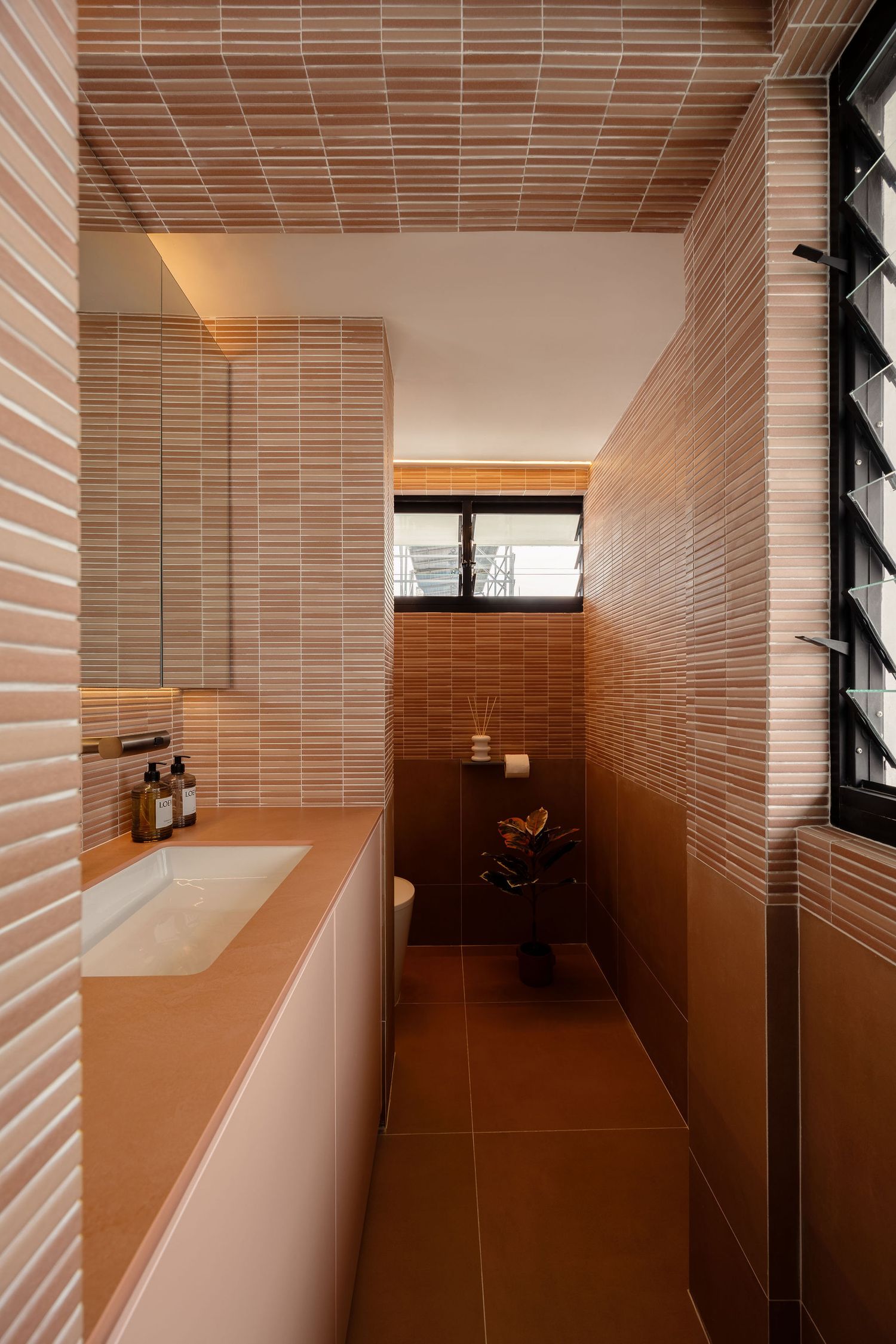 Modern narrow bathroom with terracotta brick tiles on walls and ceiling, white vanity sink, and louvered window.