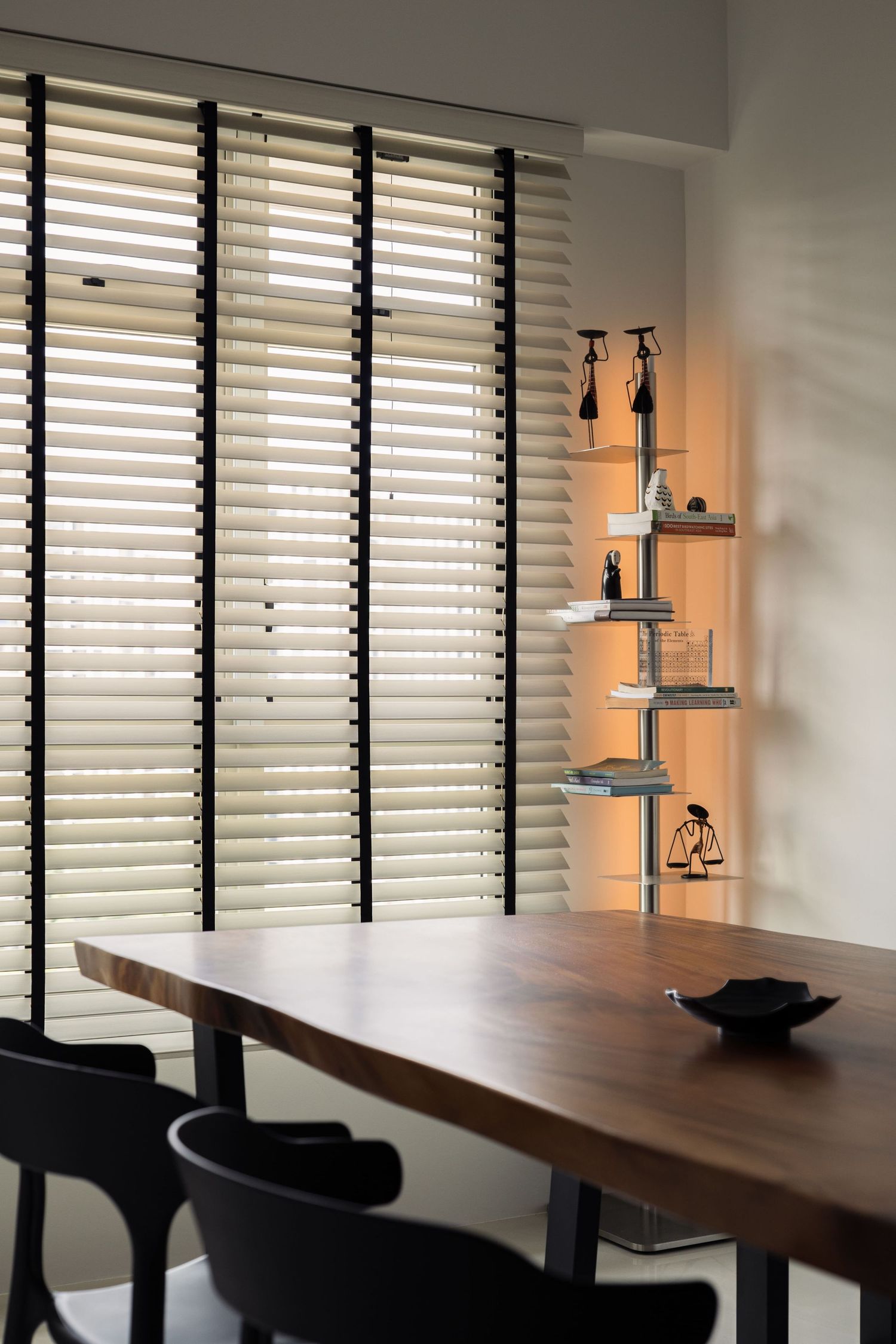 Modern dining room with wooden table, black chairs, venetian blinds, and illuminated corner shelf displaying decorative objects.