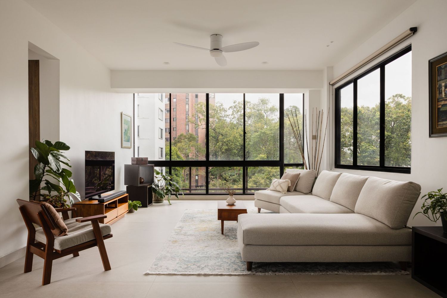 Modern living room with white sectional sofa, black-framed floor-to-ceiling windows, lush greenery views, and ceiling fan.