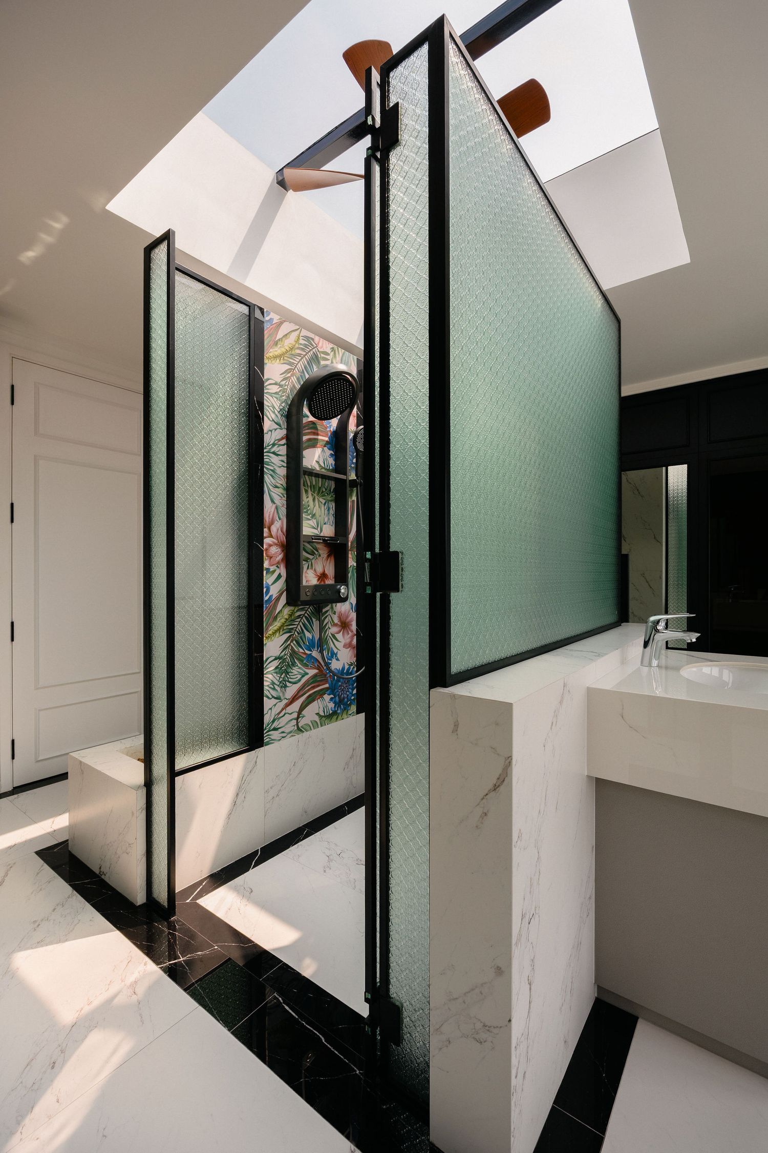 Modern bathroom with marble floors, green textured glass shower partition, floral tropical wallpaper, skylight and a floating marble vanity.