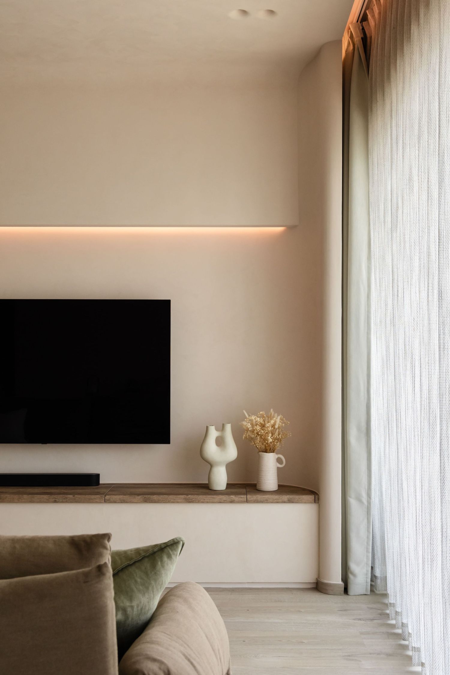 Modern minimalist living room with wall-mounted TV, accent lighting, beige walls, and green velvet throw pillows on sofa.