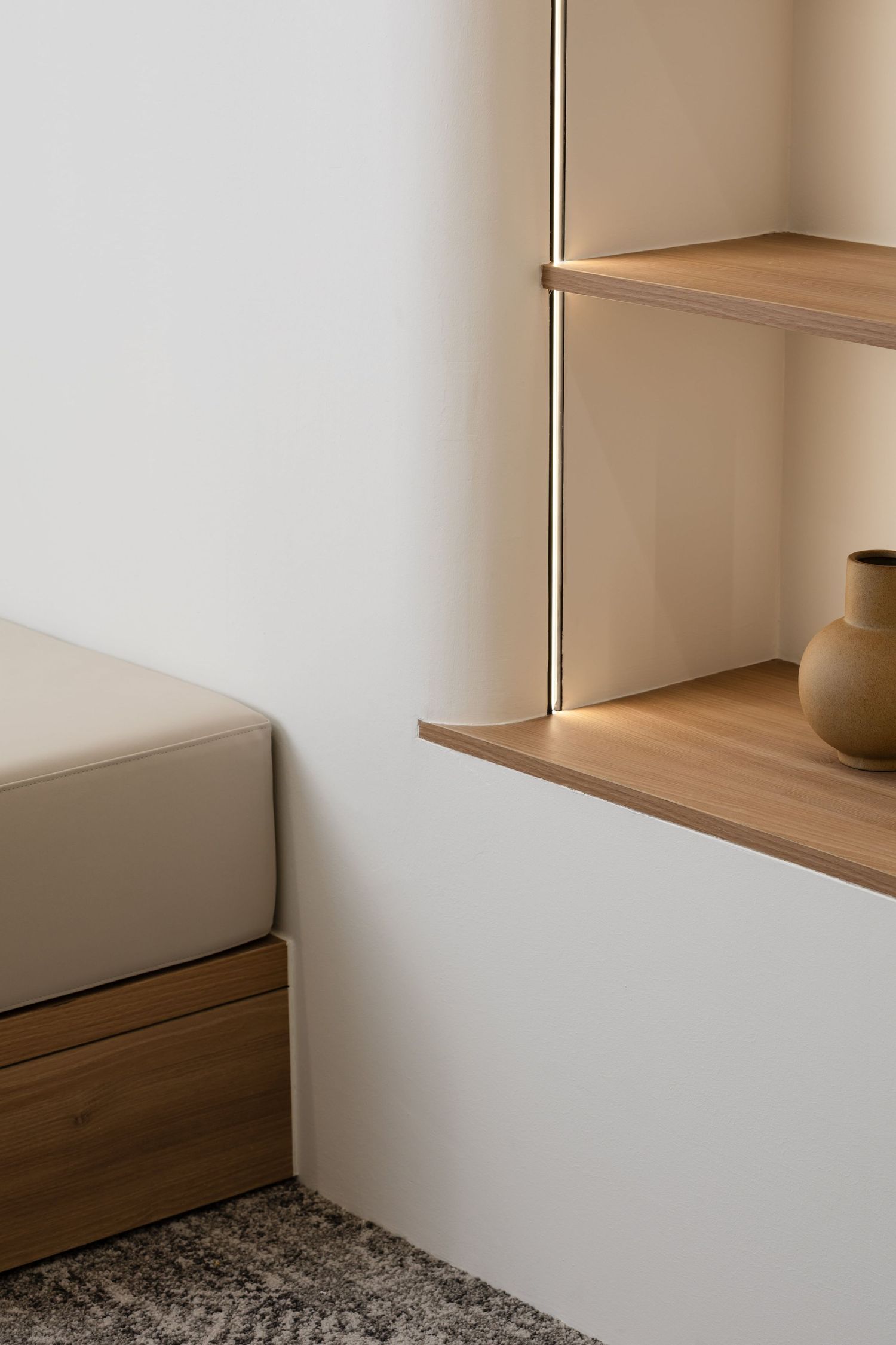 Minimalist interior corner featuring built-in wooden shelving and seating with gray cushions against white walls.