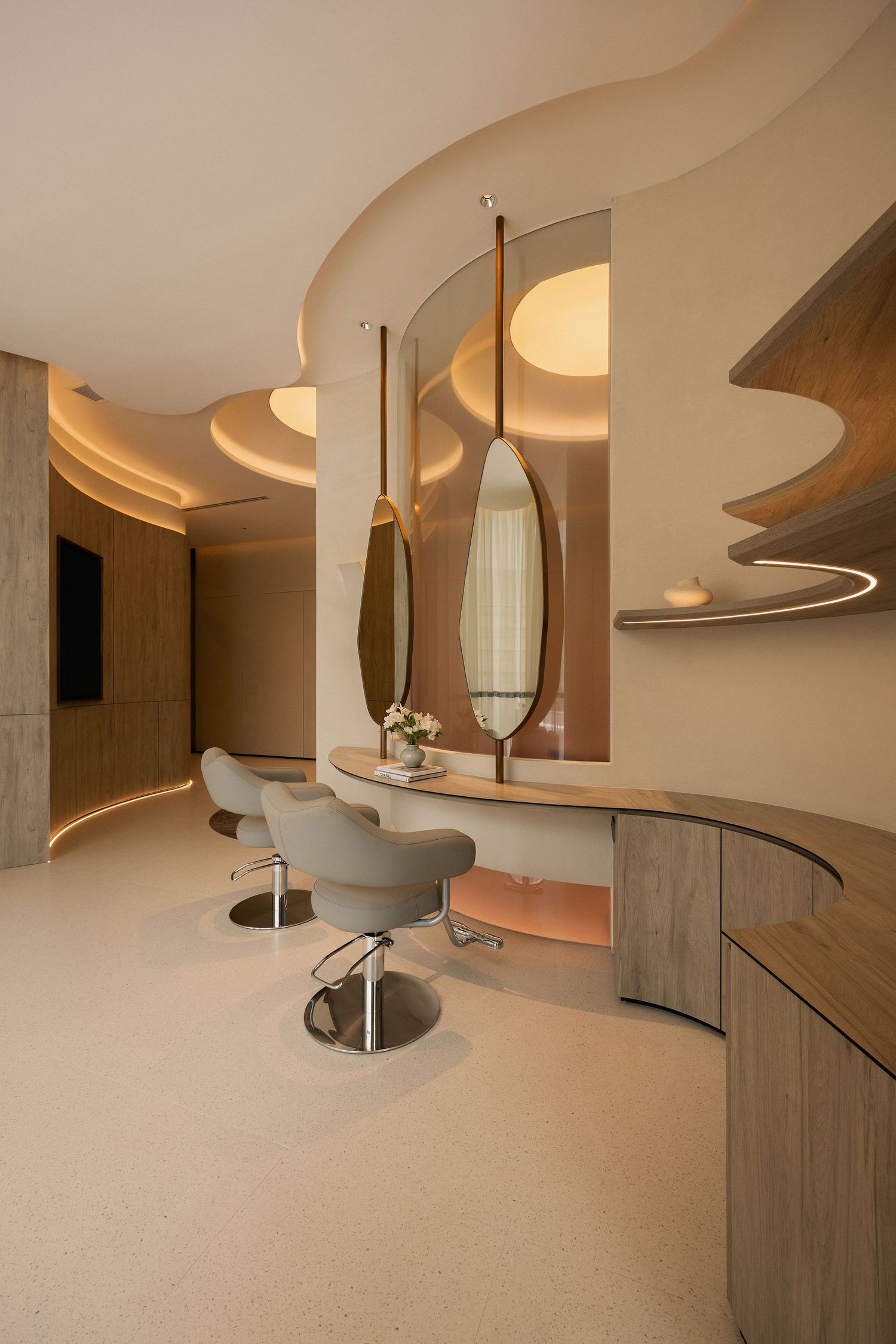 Luxurious hair salon with curved beige walls, oval mirrors, soft lighting, and modern styling chairs on terrazzo floors.