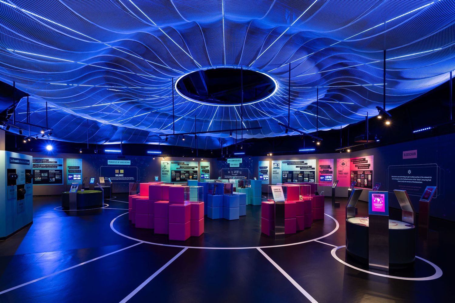 Futuristic museum exhibit hall with glowing blue ceiling installation, colorful interactive displays, and circular floor patterns.