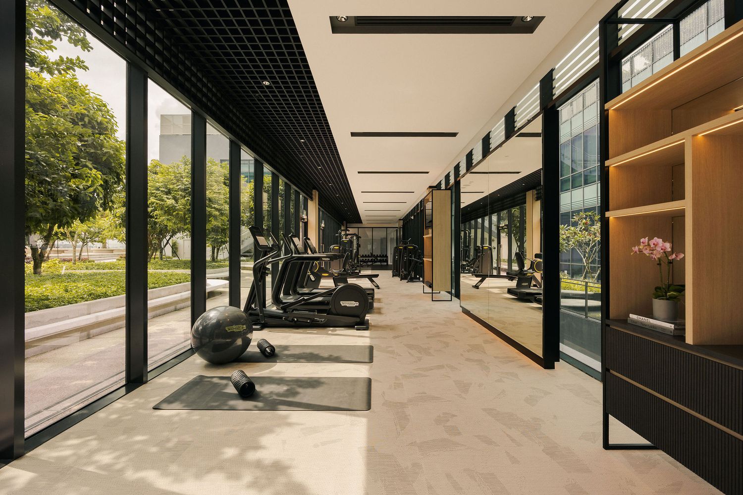 Modern luxury gym with floor-to-ceiling windows, cardio equipment, marble floors, and wood shelving overlooking lush greenery.