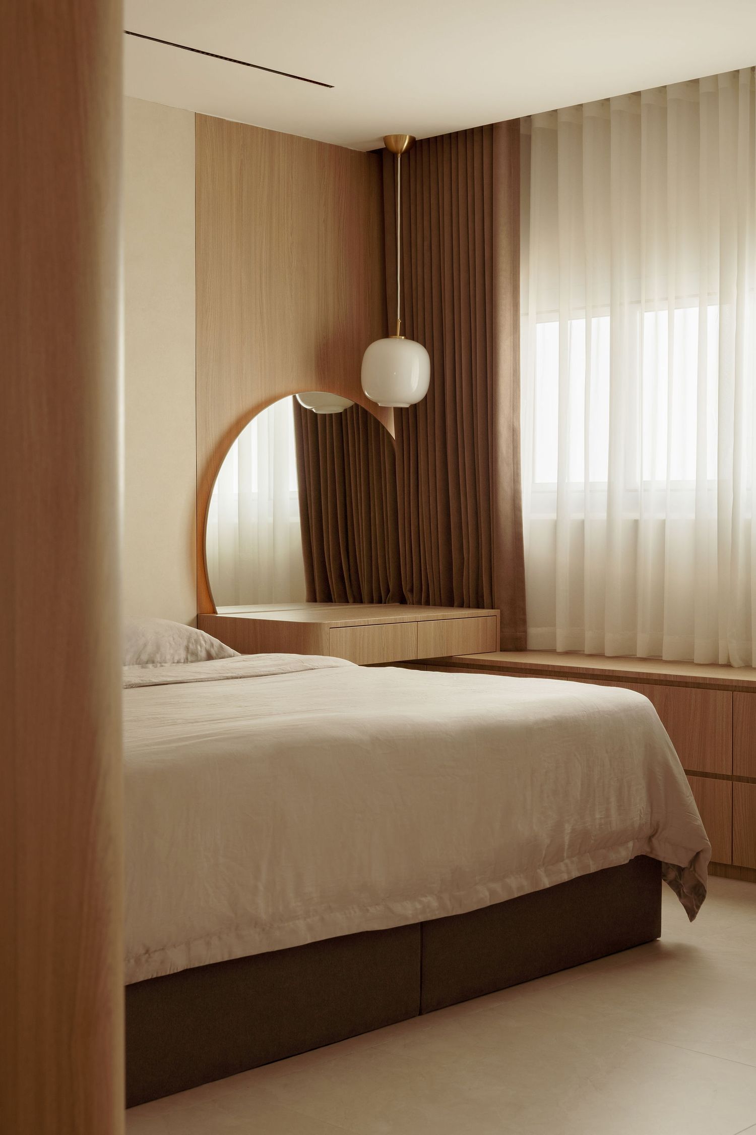 Minimalist bedroom with warm wood paneling, arched mirror, white bedding, pendant light, and sheer curtains in neutral tones.