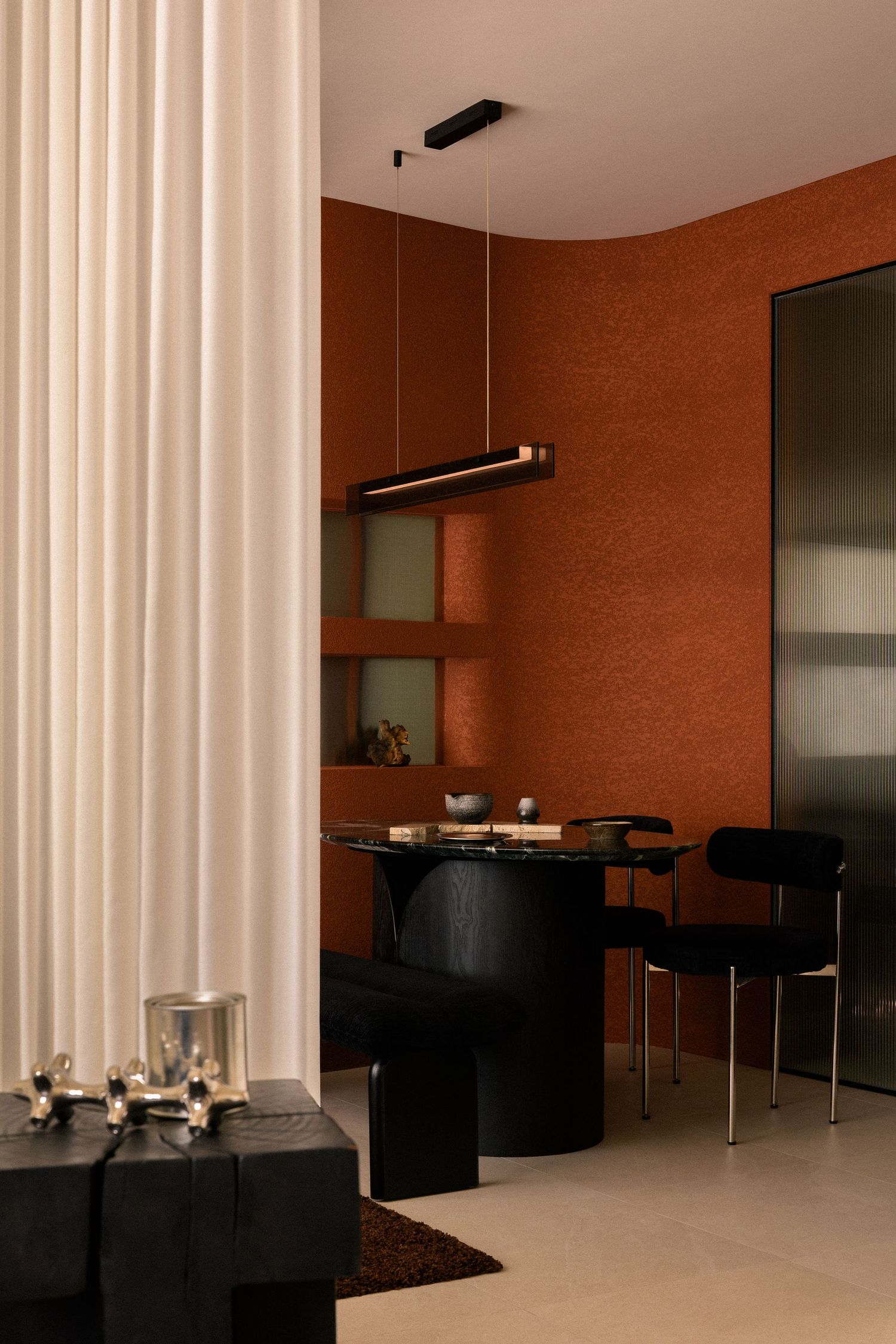 Modern dining nook with terracotta textured walls, a black round pedestal table, velvet chairs, linear pendant light and cream curtain.