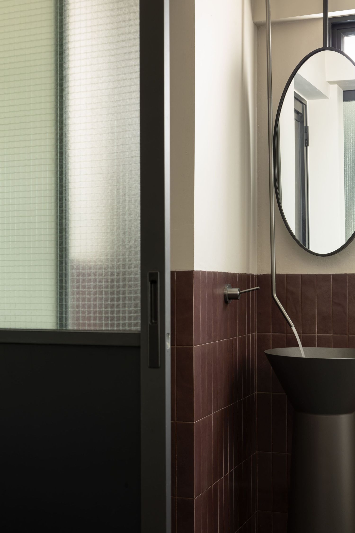 Modern bathroom interior with textured glass partition, round mirror, and burgundy tile wall accents.