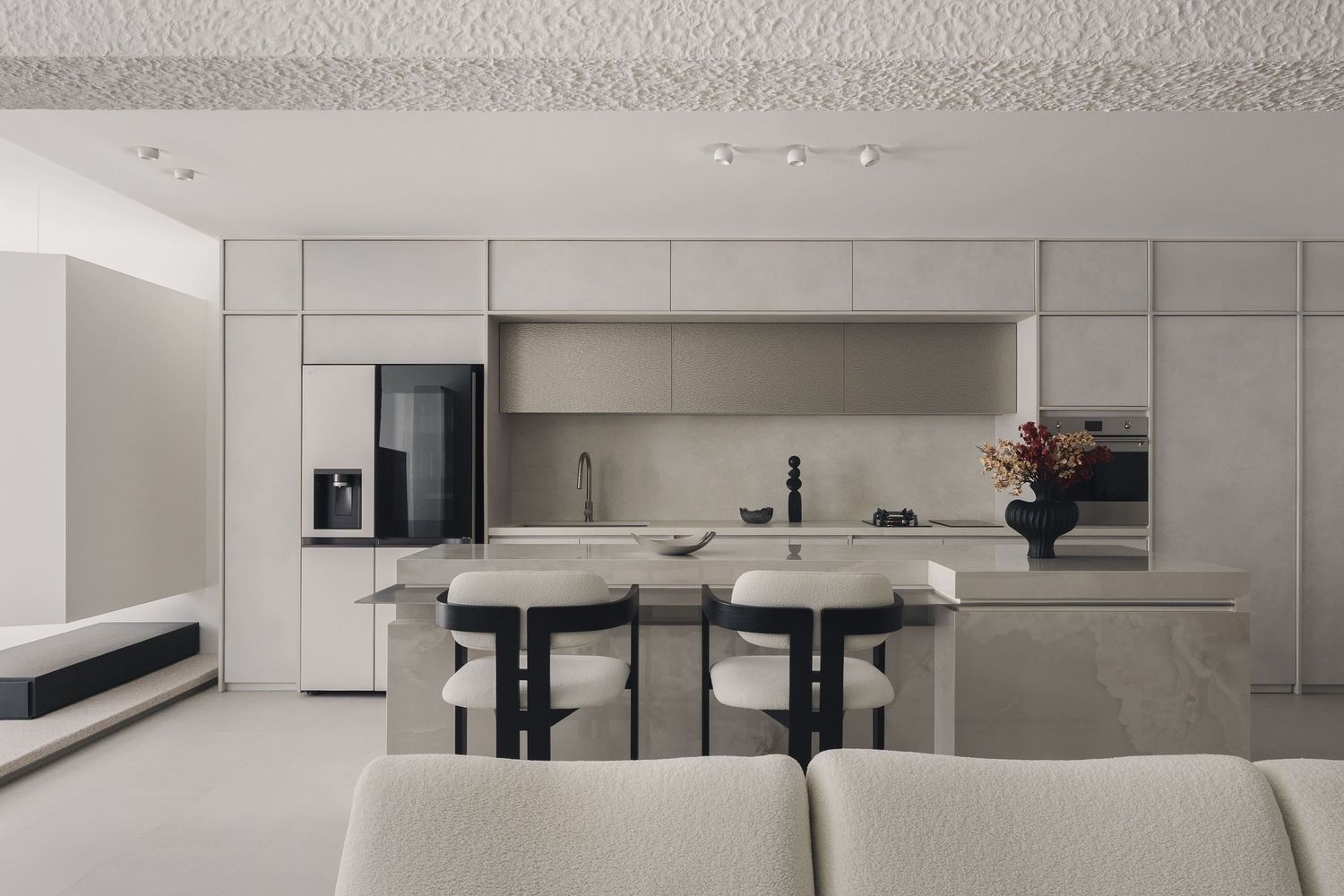 Modern minimalist kitchen with beige cabinetry, black bar stools, island counter, and integrated appliances in an open-plan living space.