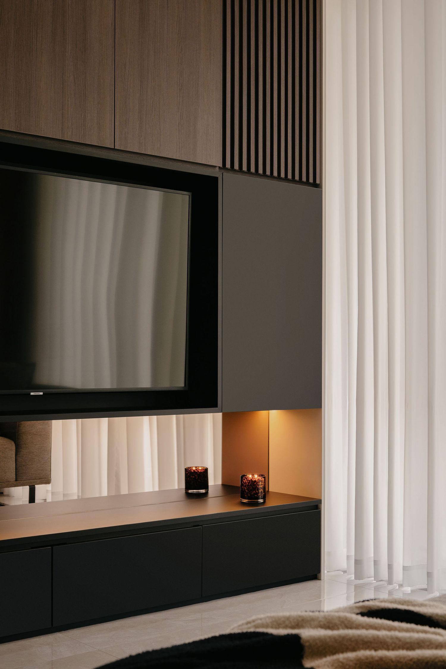 Modern living room media wall with built-in TV, dark cabinetry, wood slat paneling, warm LED lighting, candles and sheer white curtains.