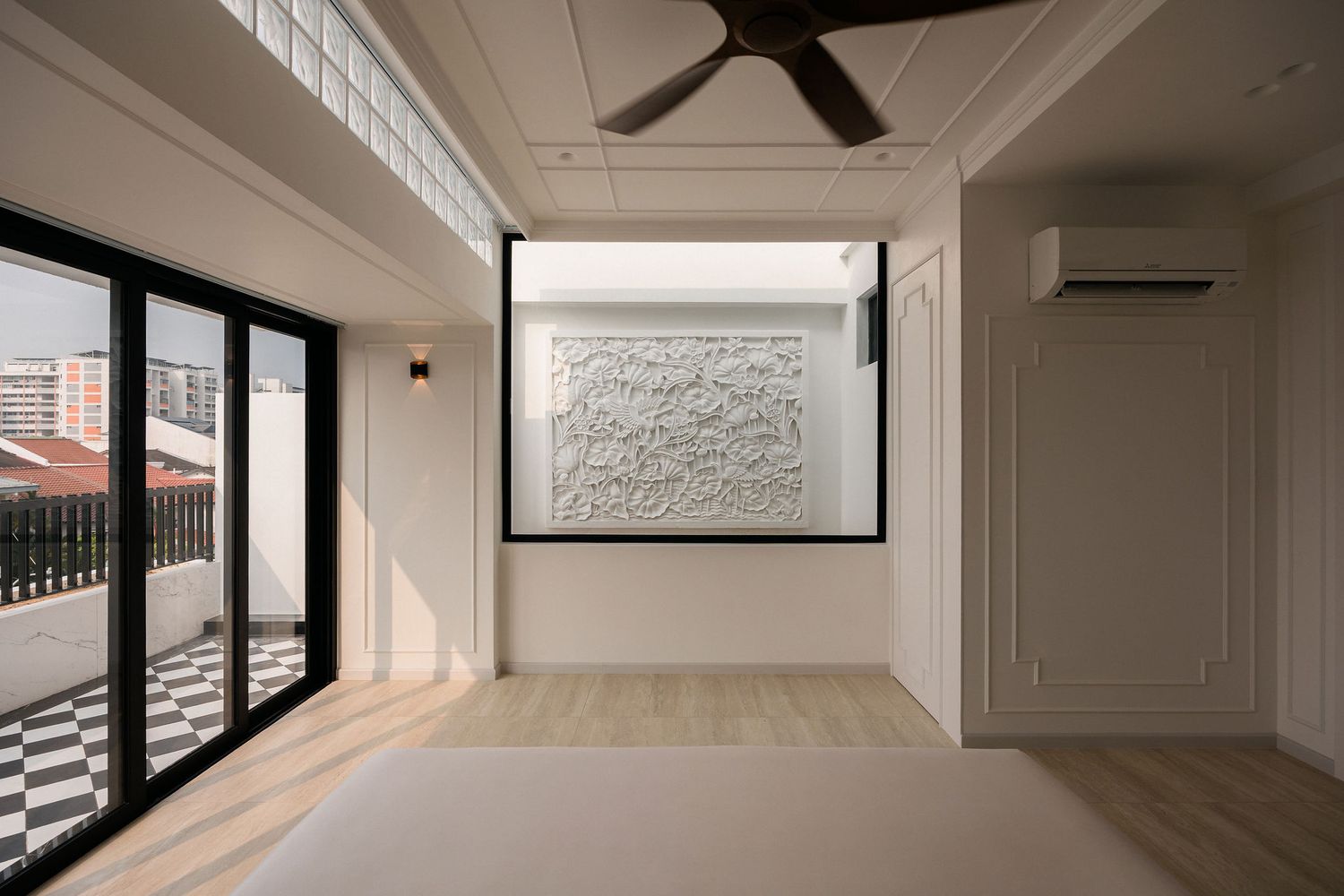 Bright empty bedroom with white paneled walls, ceiling fan, wood flooring, framed textured wall art and glass doors opening to a checkered b