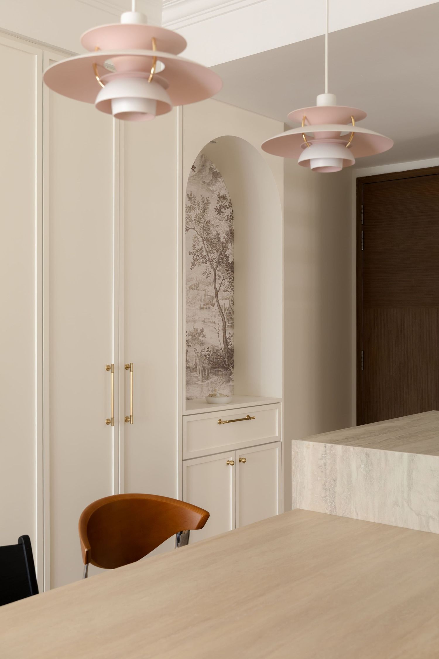Modern dining room with pink pendant lights, cream cabinetry, brass hardware, and a scenic wall mural in an arched niche.