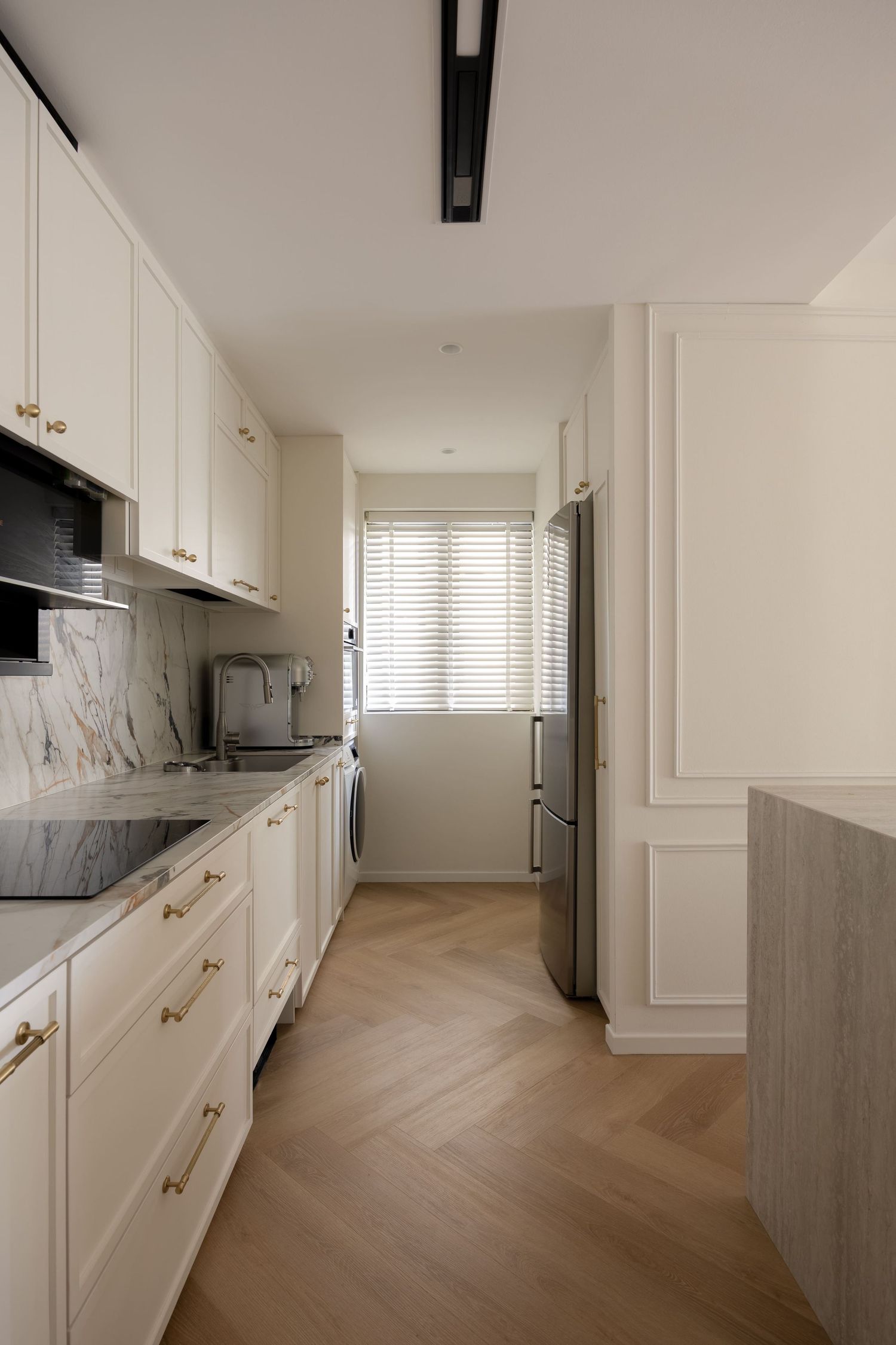 Elegant narrow kitchen with white cabinetry, gold hardware, marble backsplash, herringbone wood floors, and stainless steel appliances.