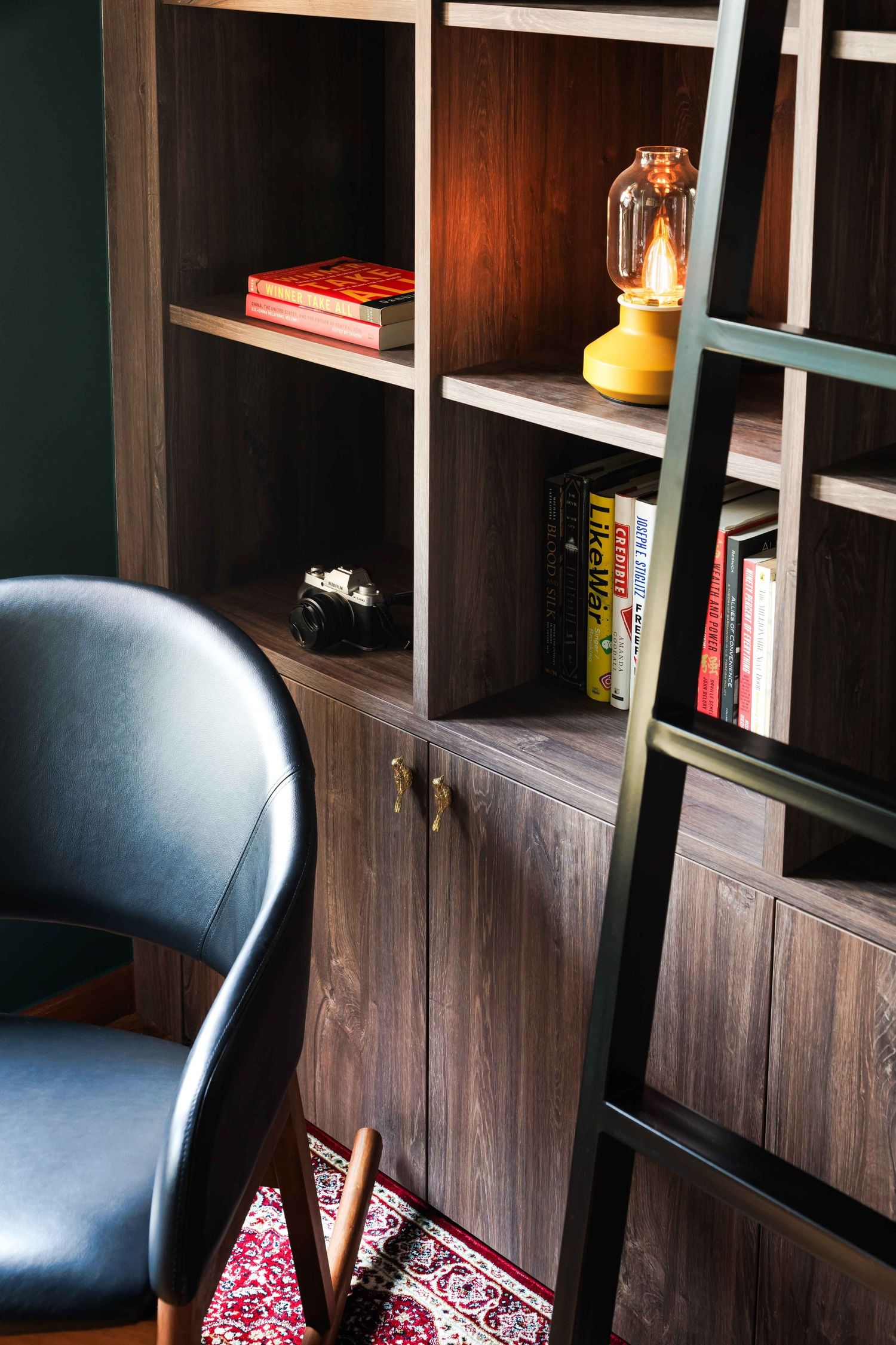 A cozy reading nook with dark wooden shelves, books, a vintage lantern, and a black leather chair on a red patterned rug.