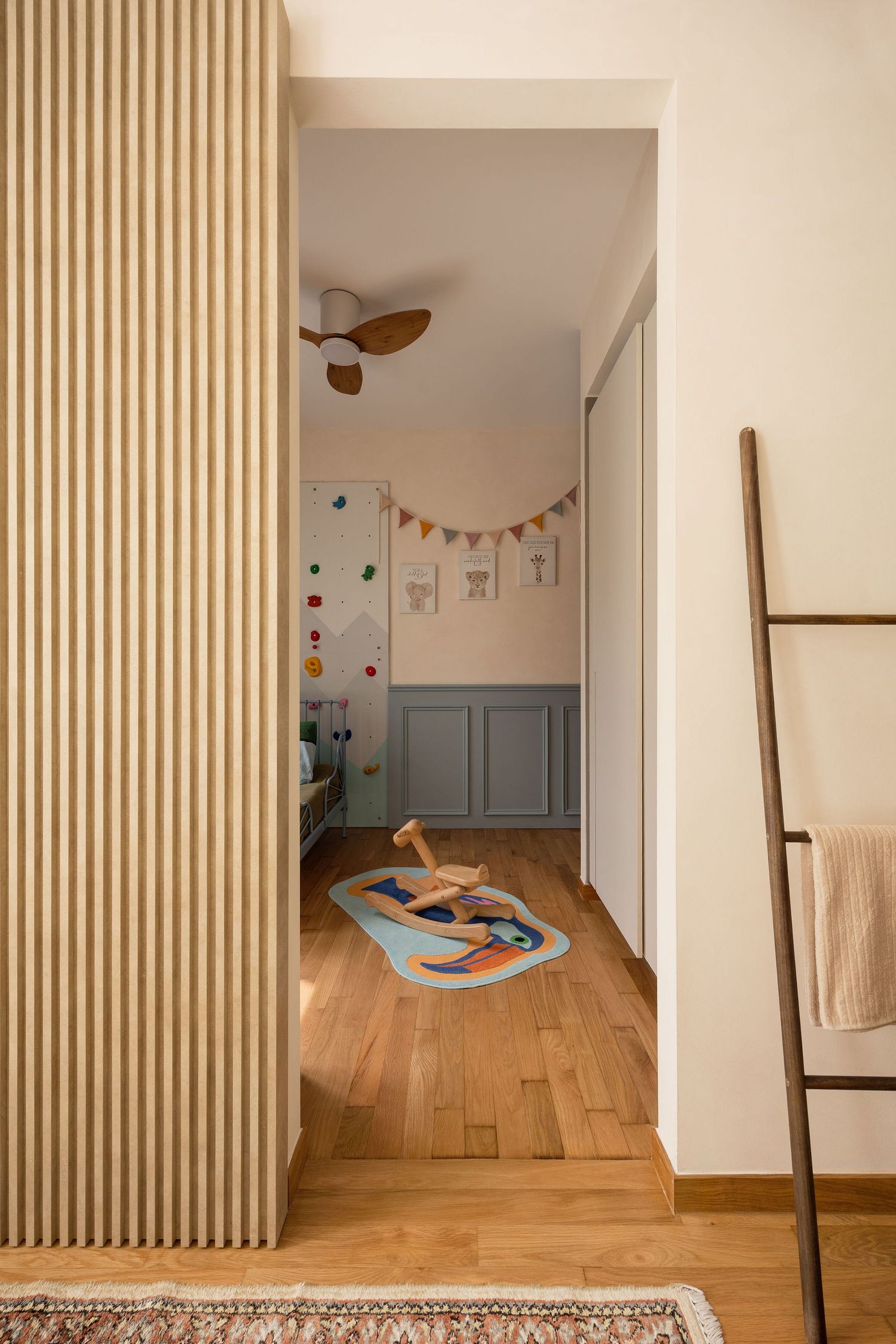 Bright children's room viewed through hallway with wood slat wall, rocking horse, climbing wall, and ceiling fan.