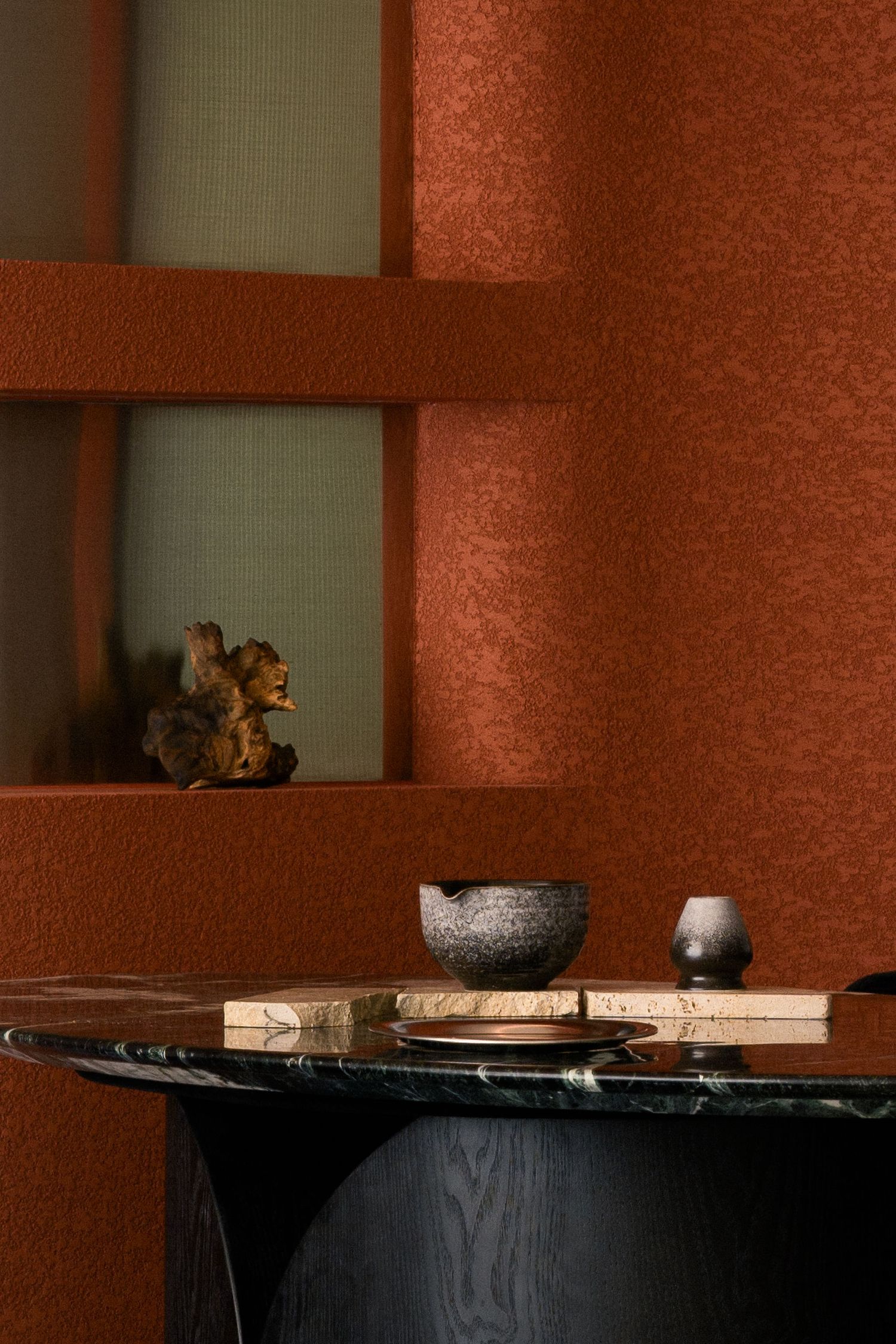 Terracotta textured walls frame a black marble table with stone bowls, a small vase, and a carved wooden sculpture in a window niche.
