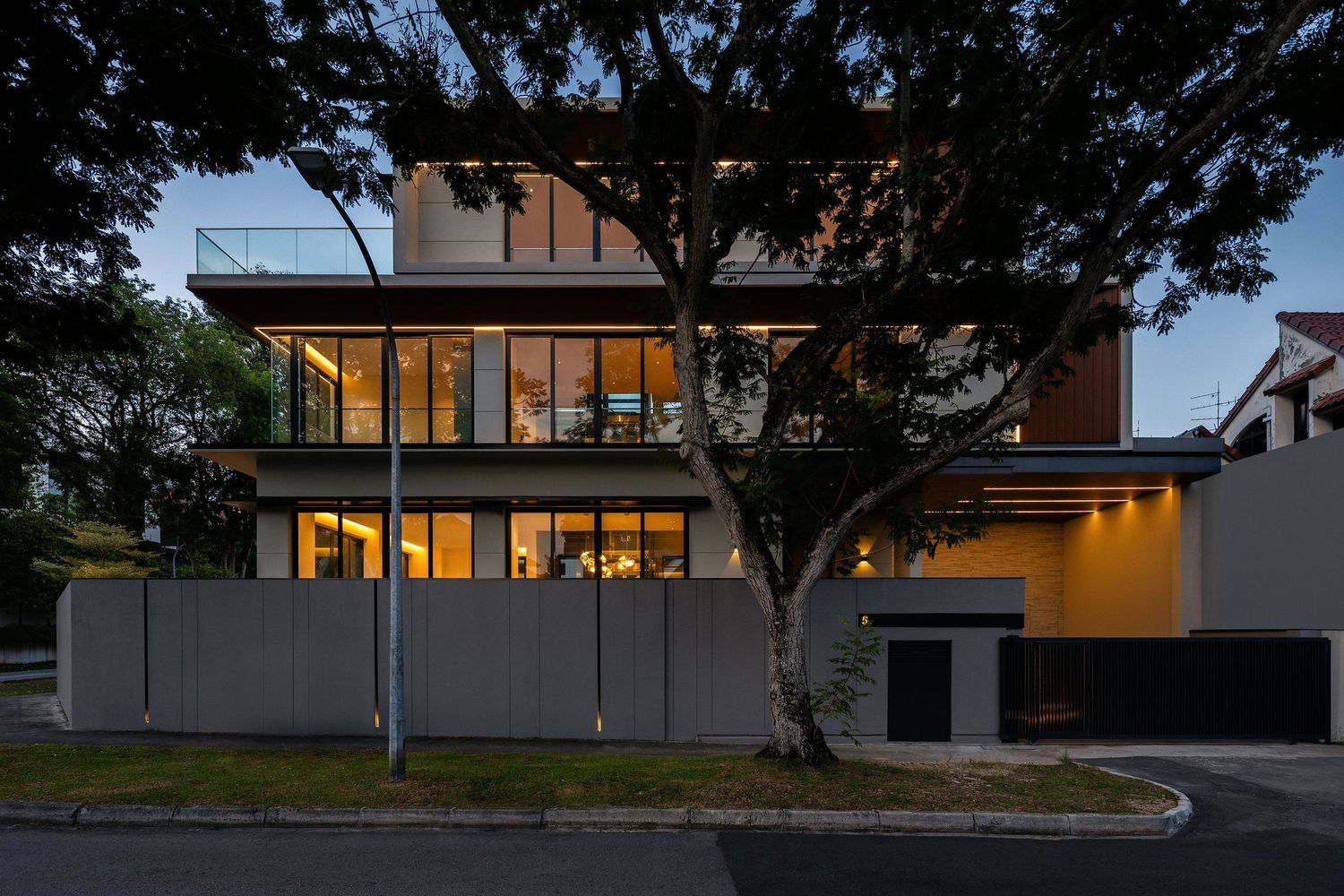 Modern multi-story house with large glass windows and warm interior lighting, framed by a mature tree and concrete fence at dusk.
