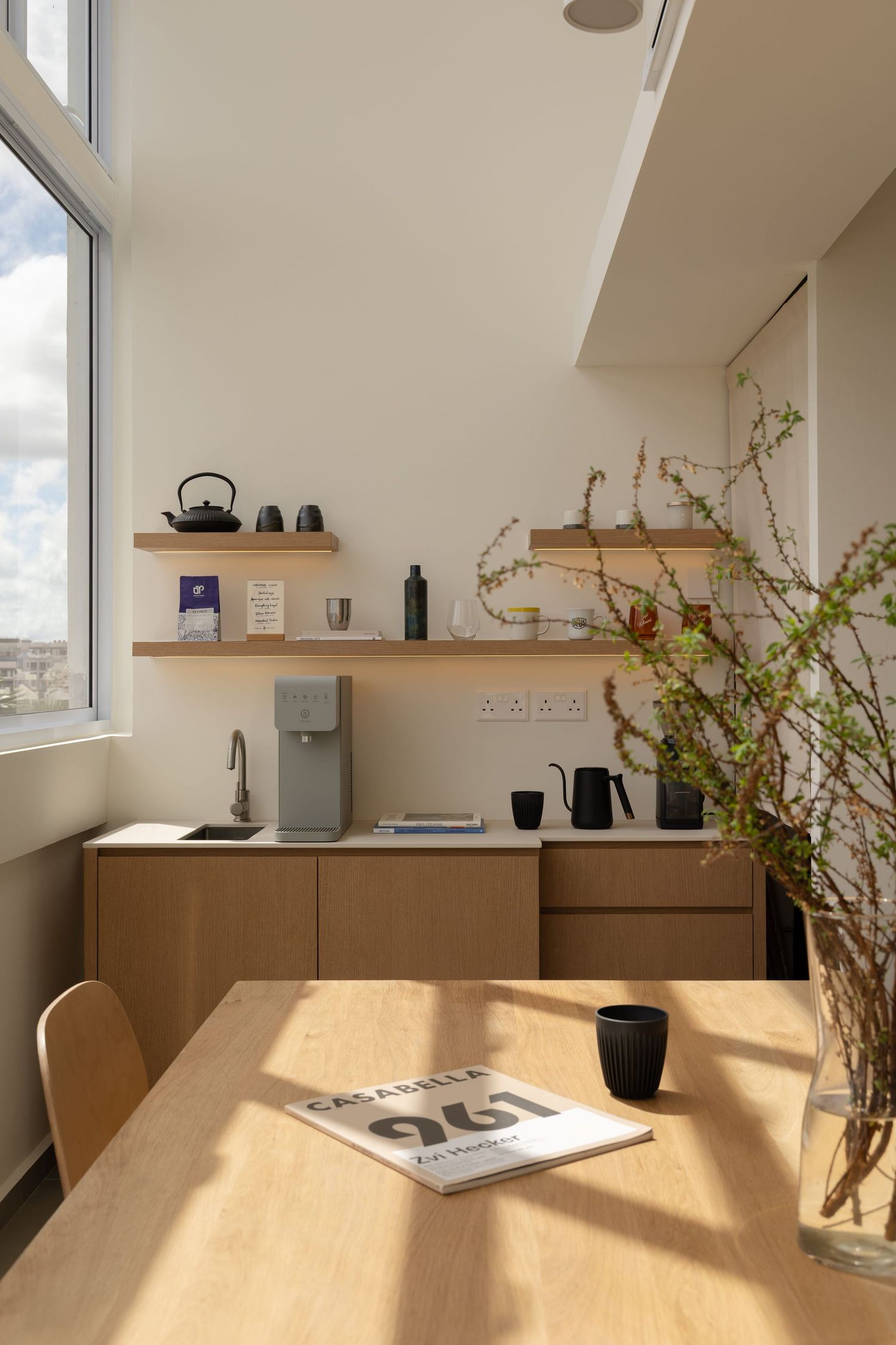 Modern minimalist kitchen corner with wooden table, floating shelves, and water dispenser in natural sunlight.