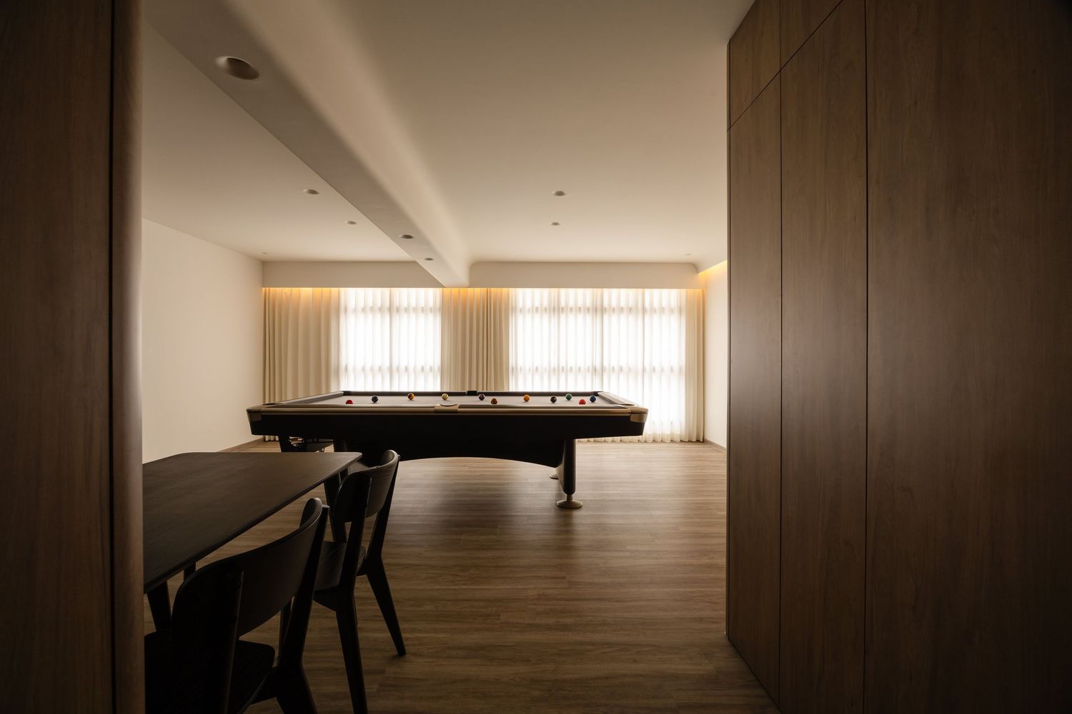 Pool table silhouetted in a minimalist room with wooden floors, dim lighting and floor-to-ceiling windows.