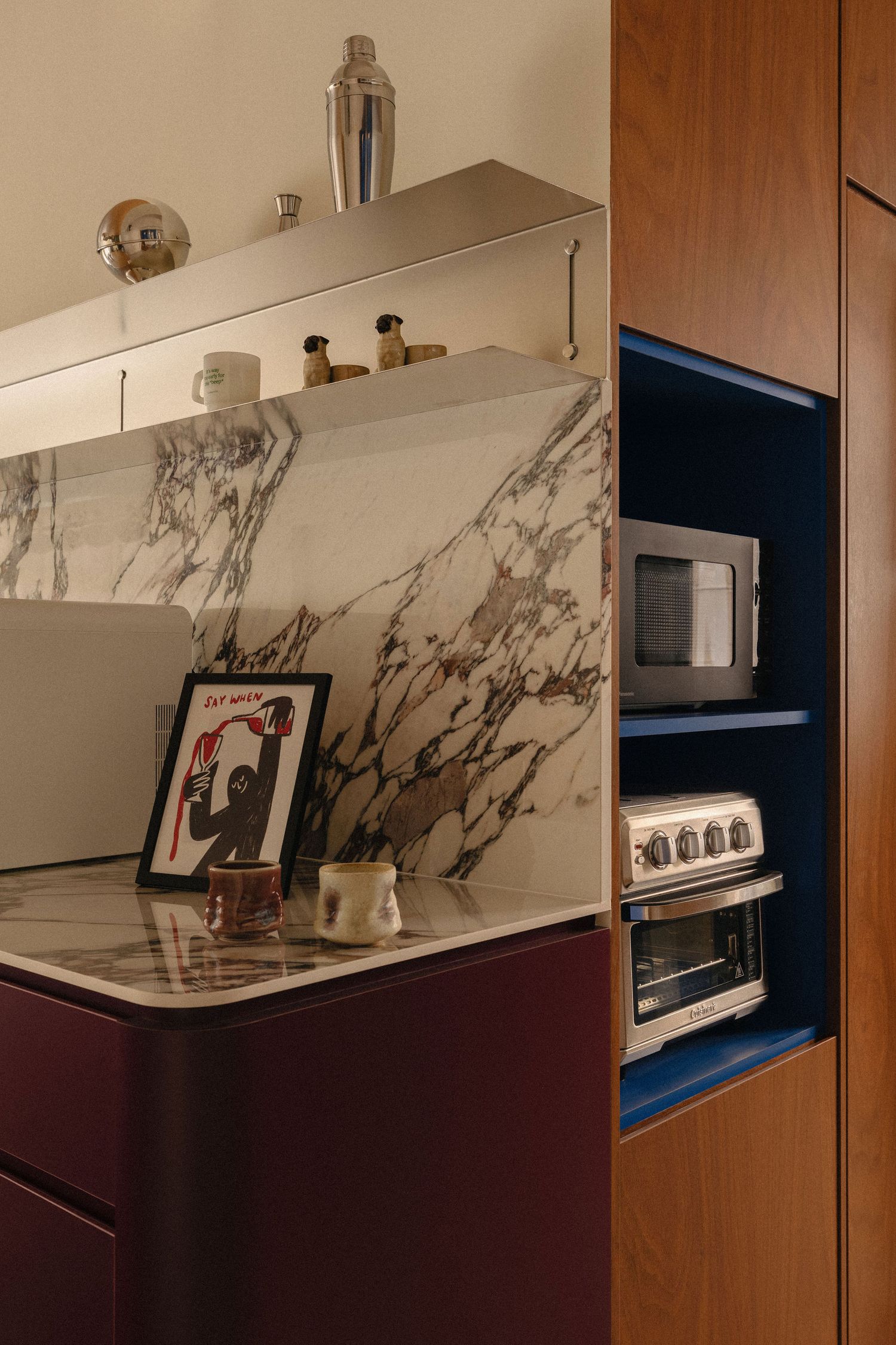Modern kitchen corner with veined marble backsplash, burgundy counter, blue-lined shelves with toaster oven and microwave, and framed art.