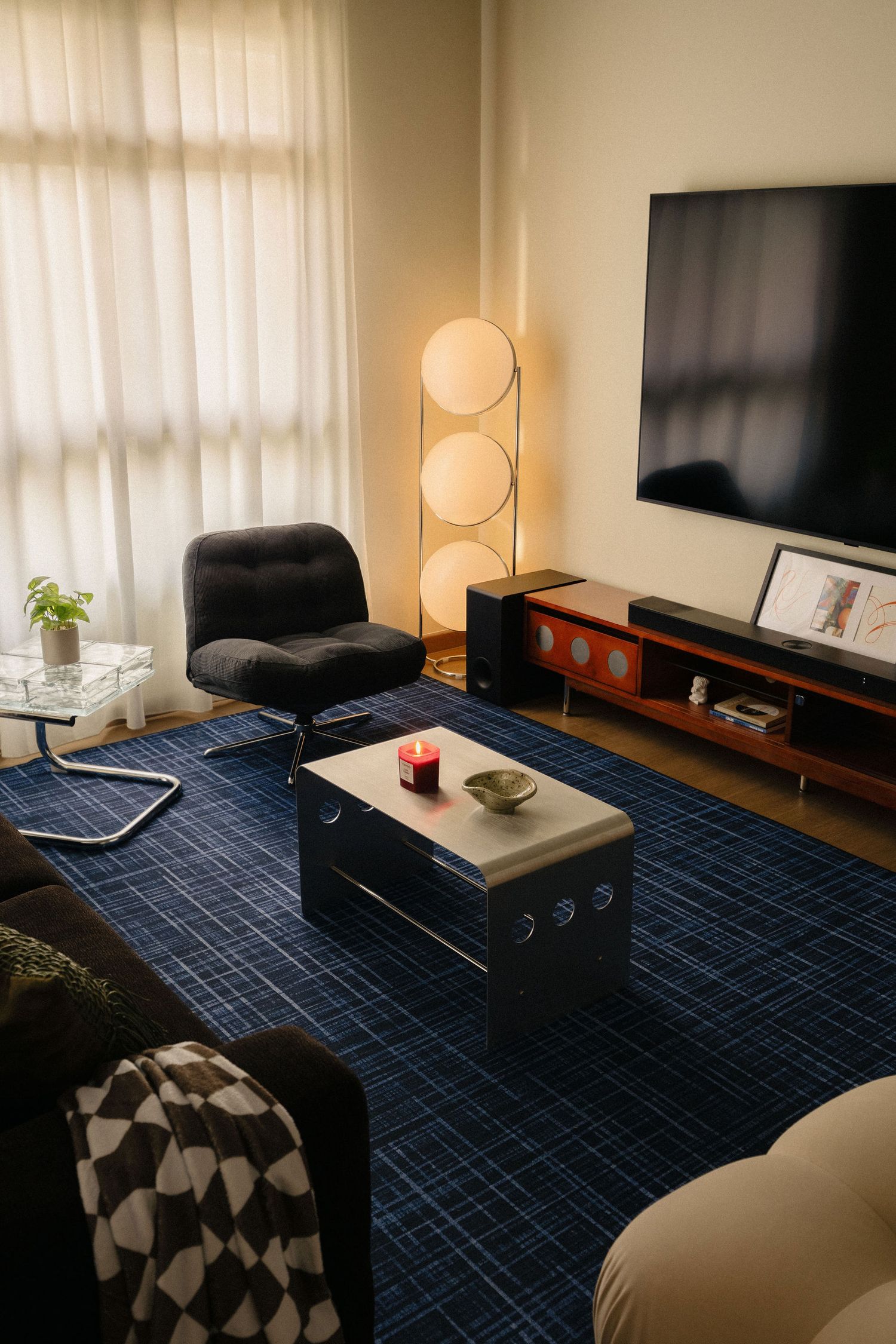 Cozy retro living room with a navy plaid rug, black tufted swivel chair, triple globe floor lamp, steel coffee table and wall-mounted TV.