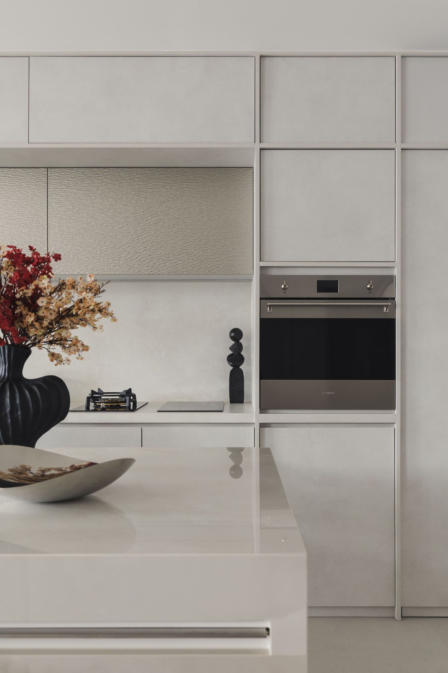Modern minimalist kitchen with white cabinetry, built-in oven, induction cooktop, and elegant floral arrangement on marble island.