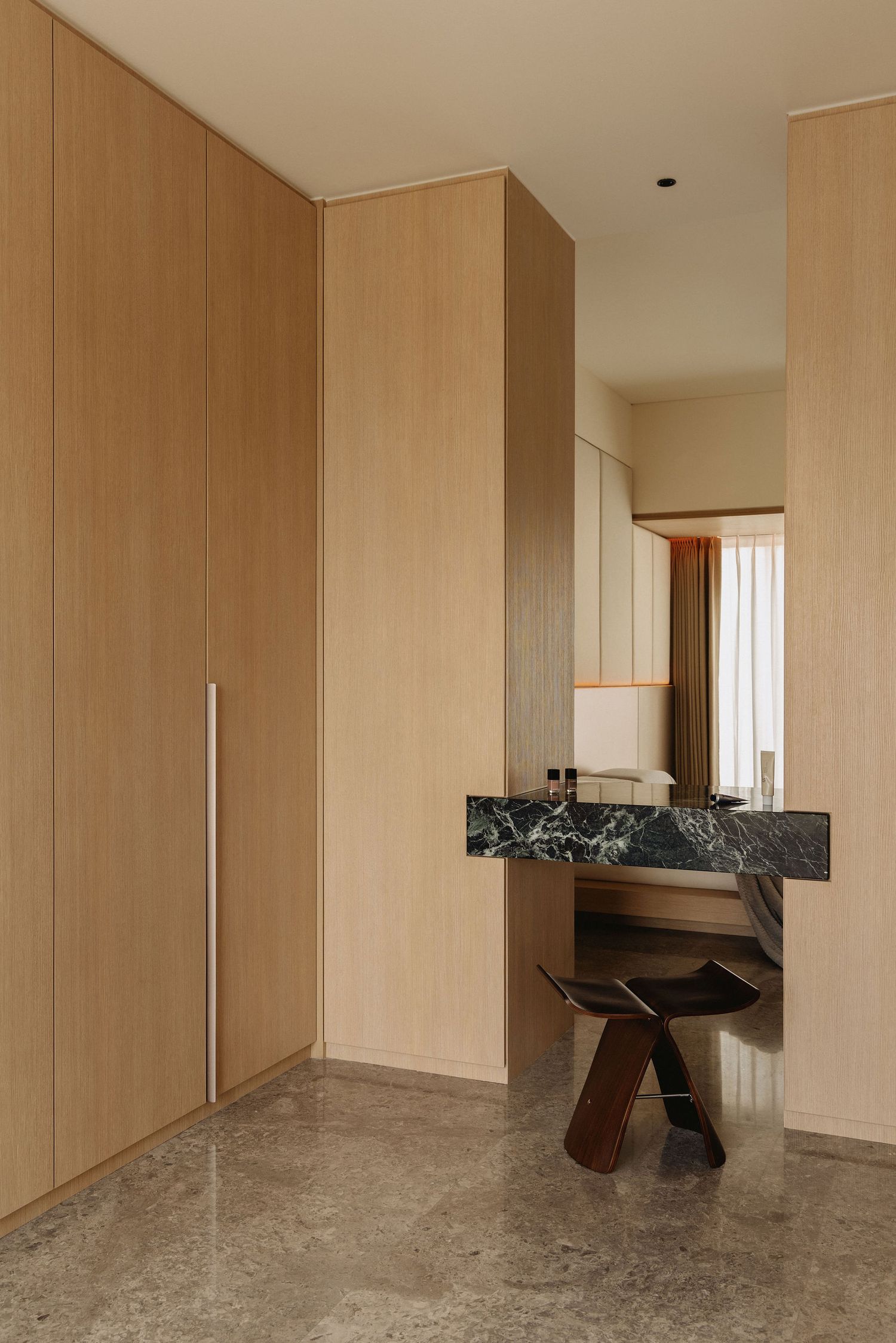 Modern hotel room entryway with light wood wardrobes, green marble vanity desk, wooden stool, and polished stone tile flooring.