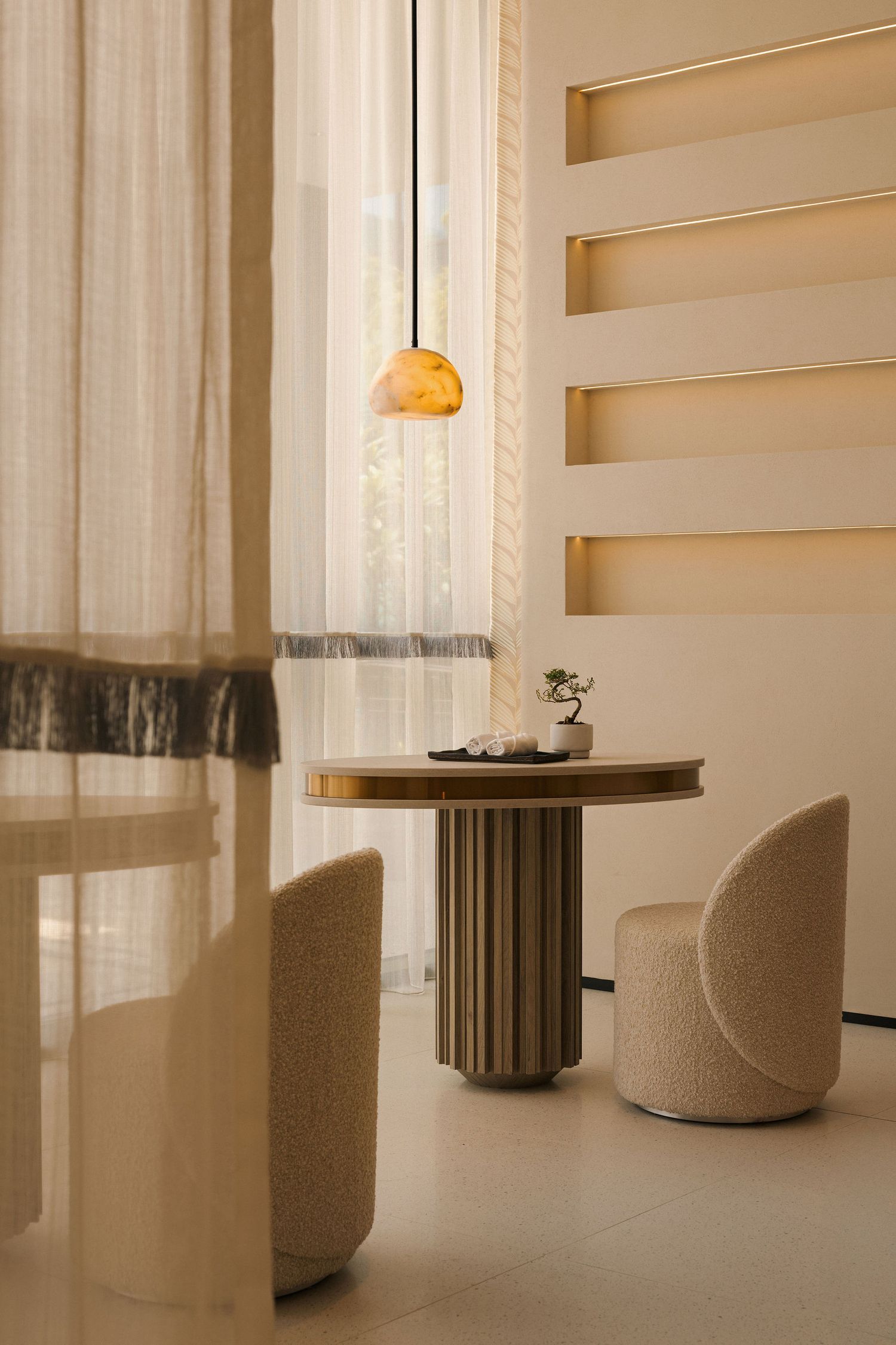 Elegant dining nook with round fluted table, curved cream chairs, amber pendant light, and sheer curtains in warm neutral tones.