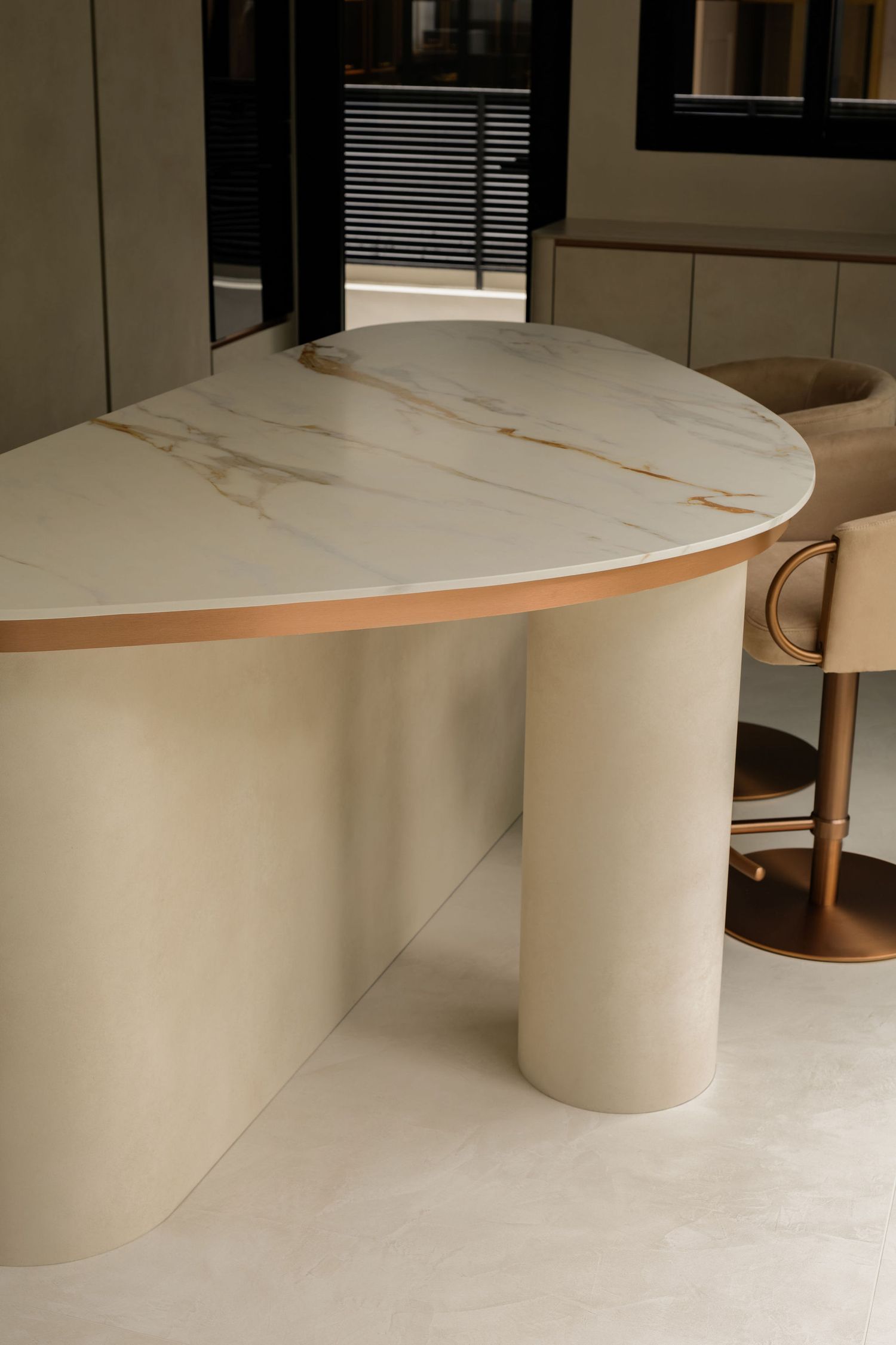Elegant curved kitchen island with white marble top, gold trim, and cream cylindrical base beside a beige bar stool.