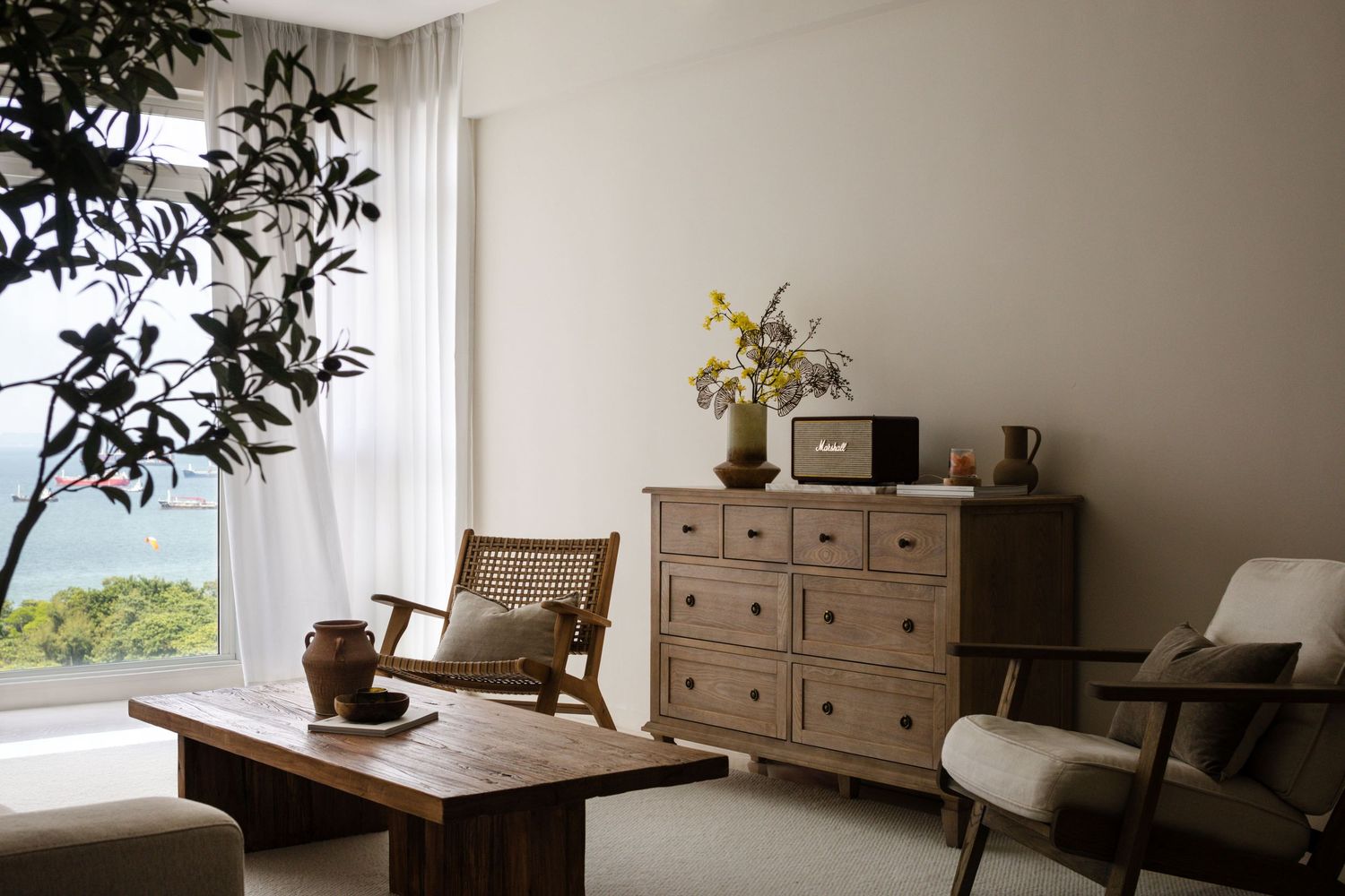 Minimalist living room with wooden dresser, coffee table, accent chair, yellow flowers, and ocean view through sheer curtains.
