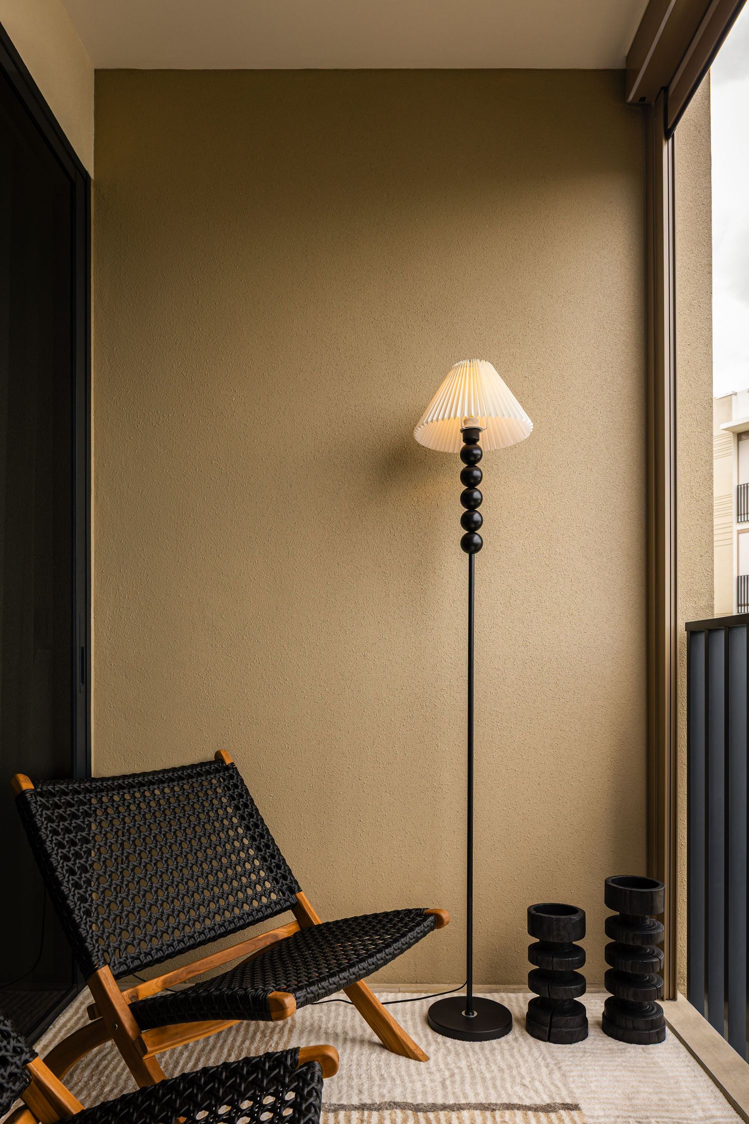 A black woven lounge chair beside a floor lamp with pleated shade and dark ceramic vases on a cozy balcony.