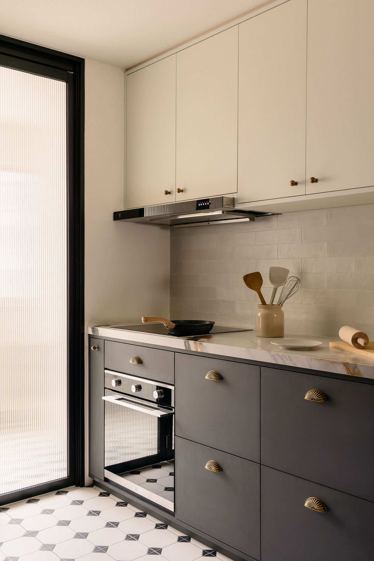 Modern kitchen with dark gray lower cabinets, cream upper cabinets, brass hardware, marble countertop, and geometric tile flooring.